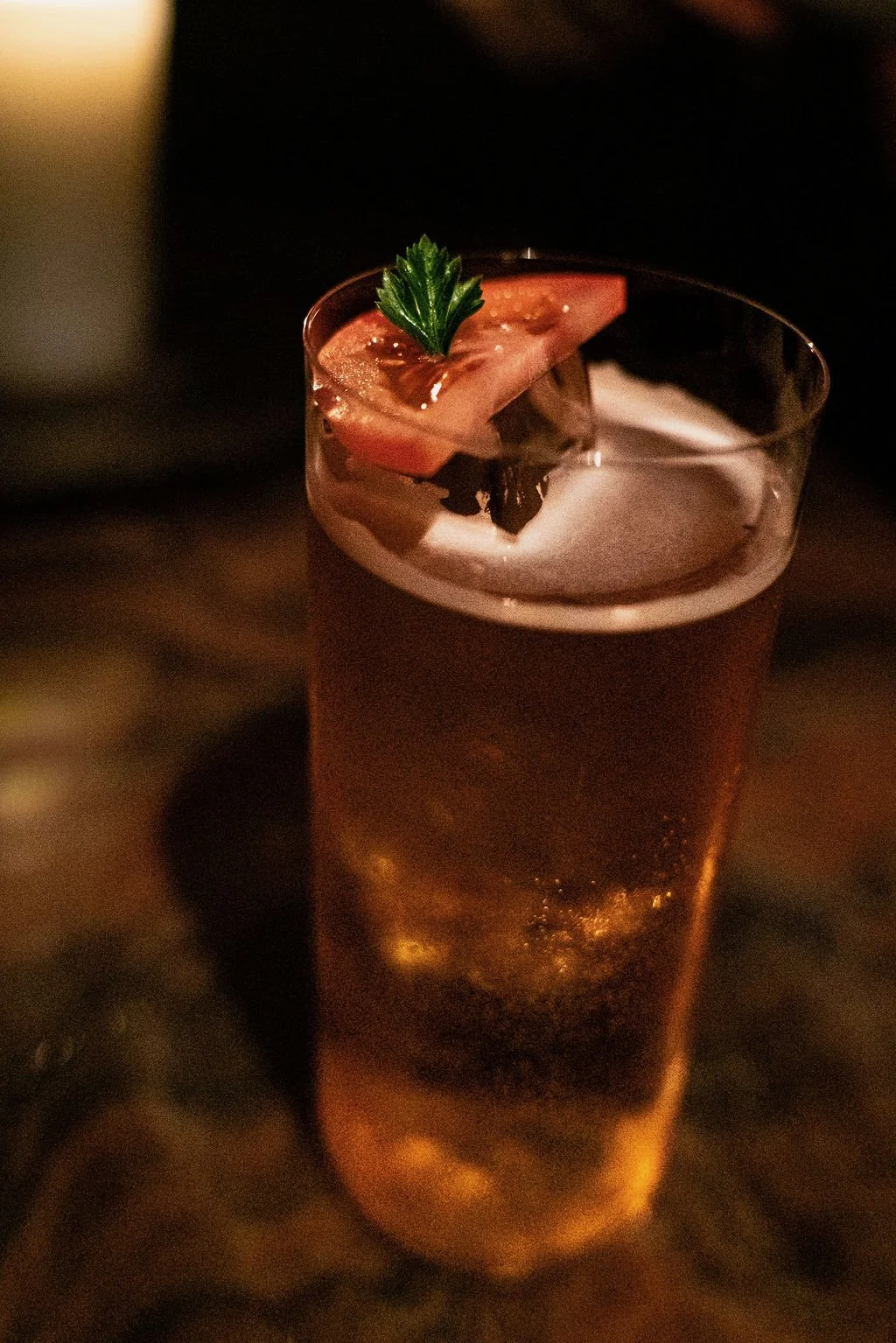 A tall glass of beer garnished with a slice of tomato and a parsley leaf, set against a dark background.