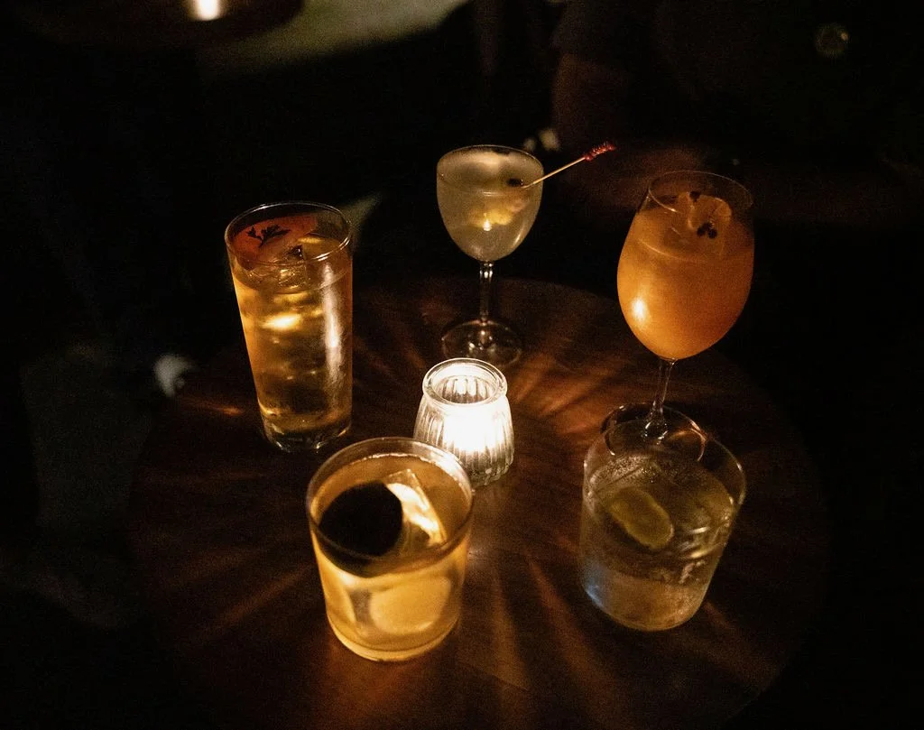 Five drinks on a dark wooden table, illuminated by a small candle, with a person partially visible in the background.