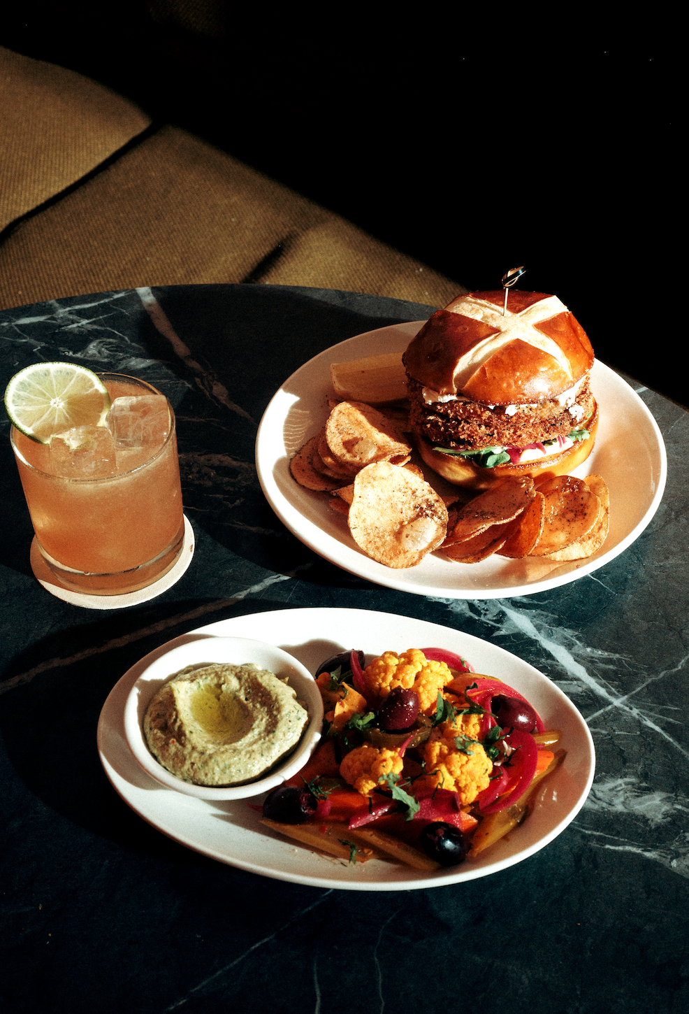 A dining table with a cocktail, a burger with potato chips, and a plate of hummus and vegetable salad.