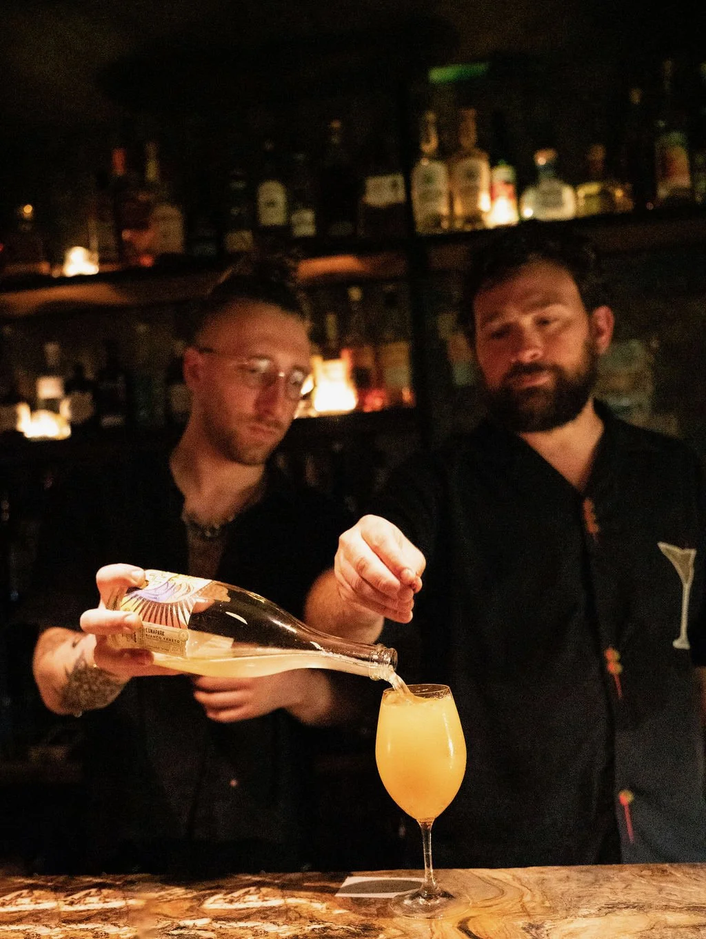 Two bartenders in a bar pouring a yellow-orange cocktail from a bottle into a glass. Shelves with bottles of alcohol in the background.
