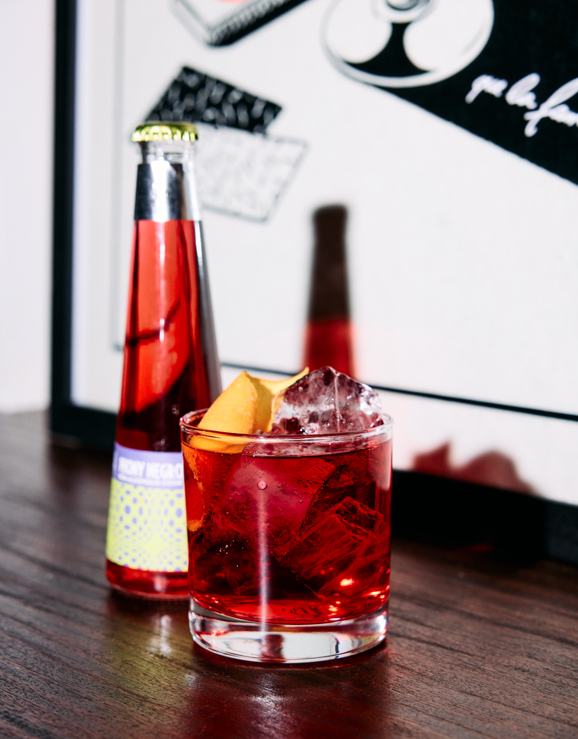 A glass of red cocktail with ice and orange slices, a bottle of red beverage, and a framed quote on a wooden table.
