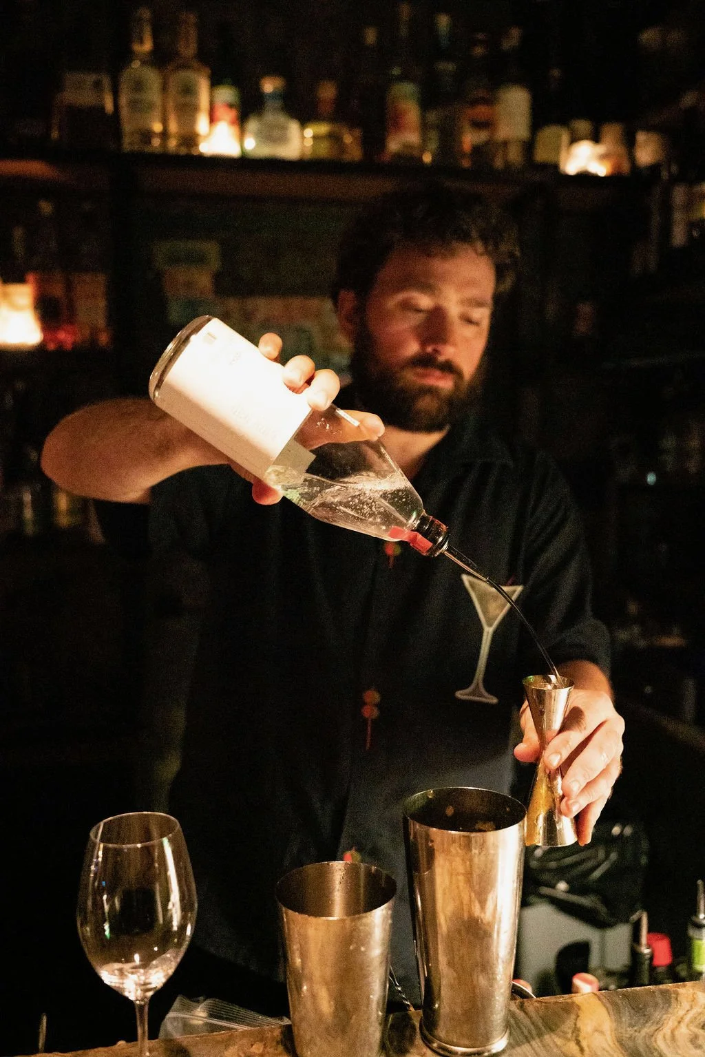 Bartender pouring clear liquid into a shaker at a dimly lit bar, with bottles on shelves in the background.