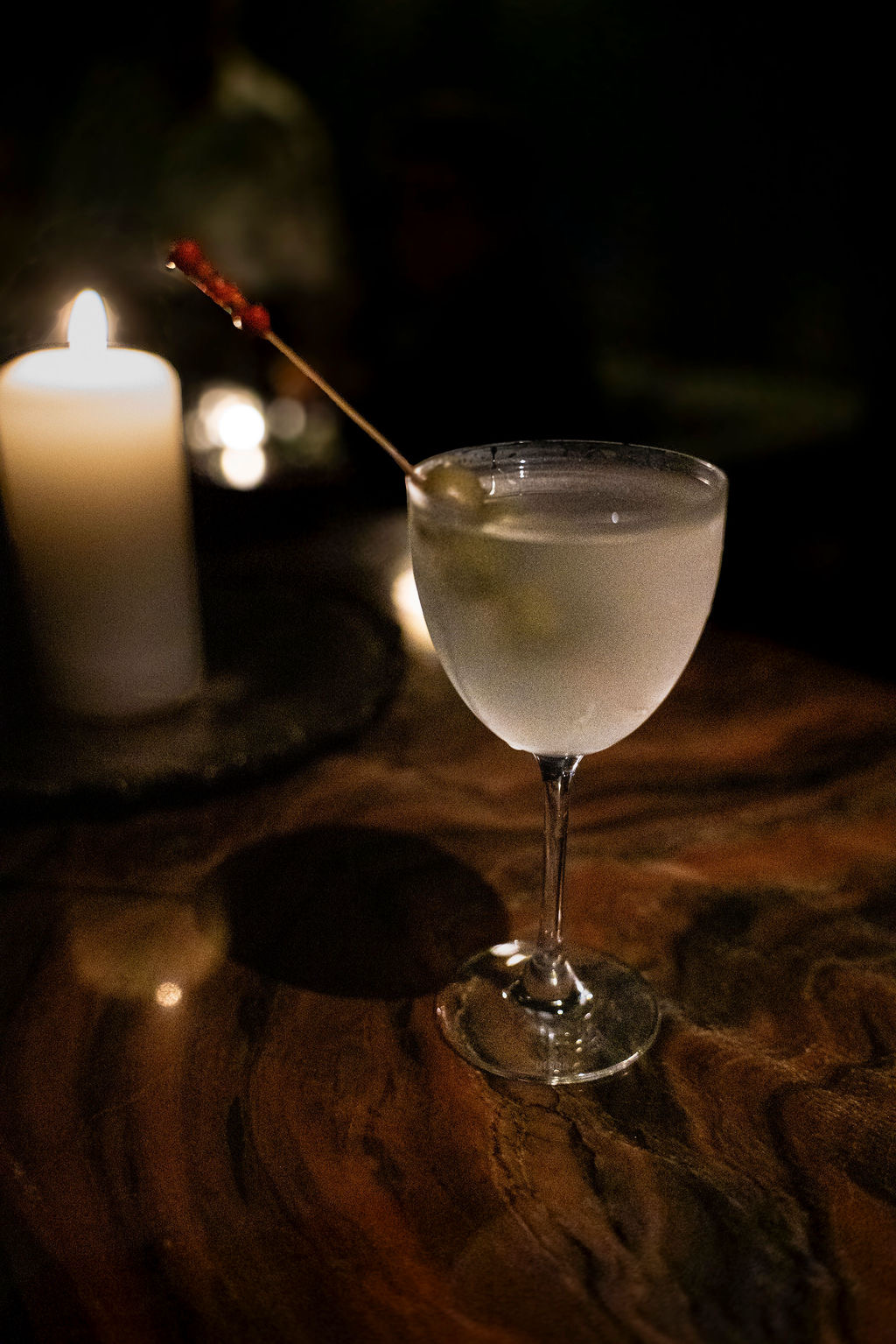 A cocktail in a coupe glass with a lime wedge, illuminated by candlelight against a dark background.