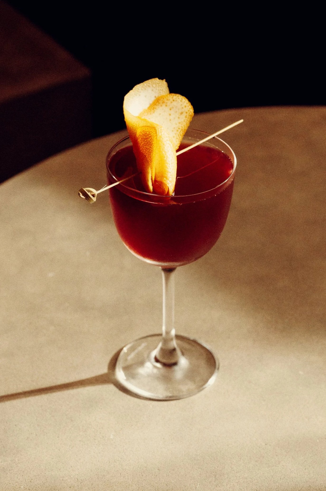 A cocktail glass filled with a dark red beverage, garnished with an orange peel and a lemon peel, with a cocktail pick resting across the top.