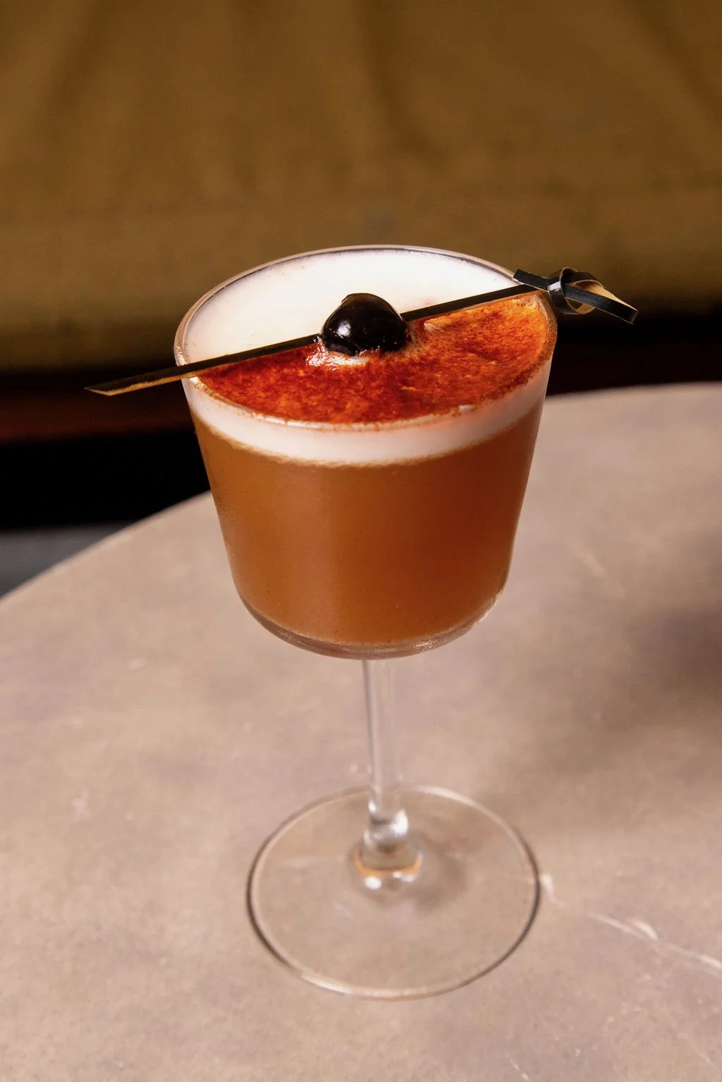 A cocktail in a stemmed glass topped with whipped cream, cocoa powder, and a black cherry, garnished with a black pick.