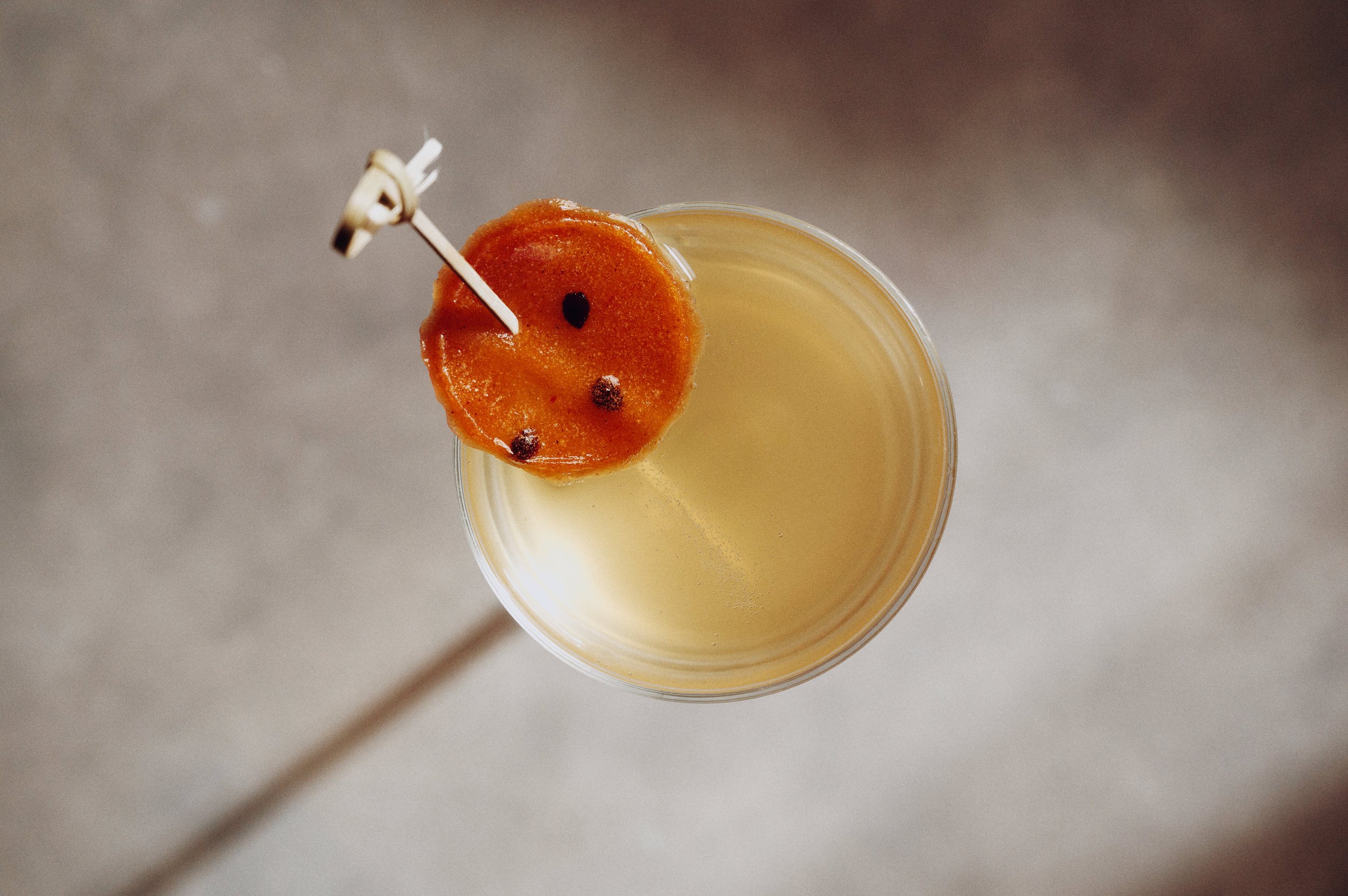 A cocktail in a clear glass with a carrot slice garnished with black peppercorns on a toothpick.