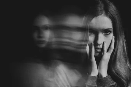 A woman with long hair holding her face with both hands, looking at the camera with a serious expression, with a blurred, ghost-like figure beside her.