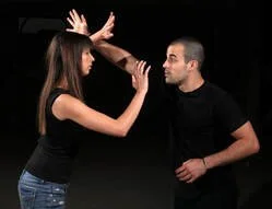 A woman and a man are engaged in a playful sword fight using wooden sticks, standing against a black background.