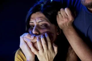 A woman appears frightened and is covering her mouth with her hands, while a person nearby is gripping her hair.