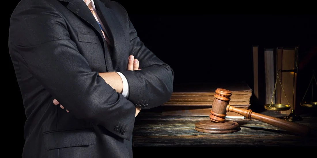 A judge's gavel resting on a wooden block, with law books, a balance scale, and a person in a suit with crossed arms in the background.