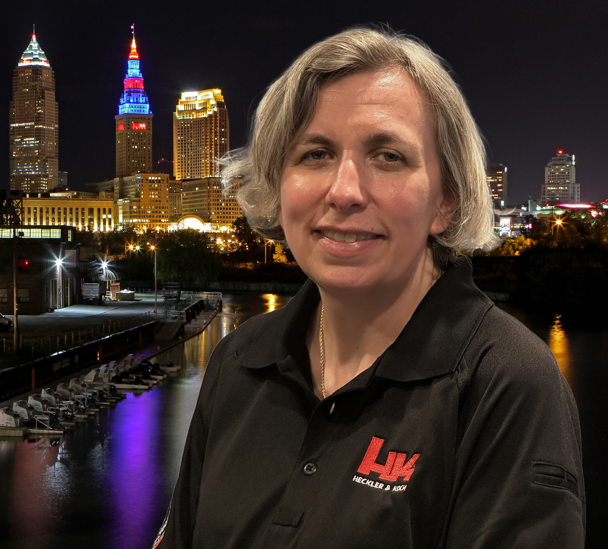 A woman with blond hair smiling in front of a city skyline at night with illuminated buildings and a river below.