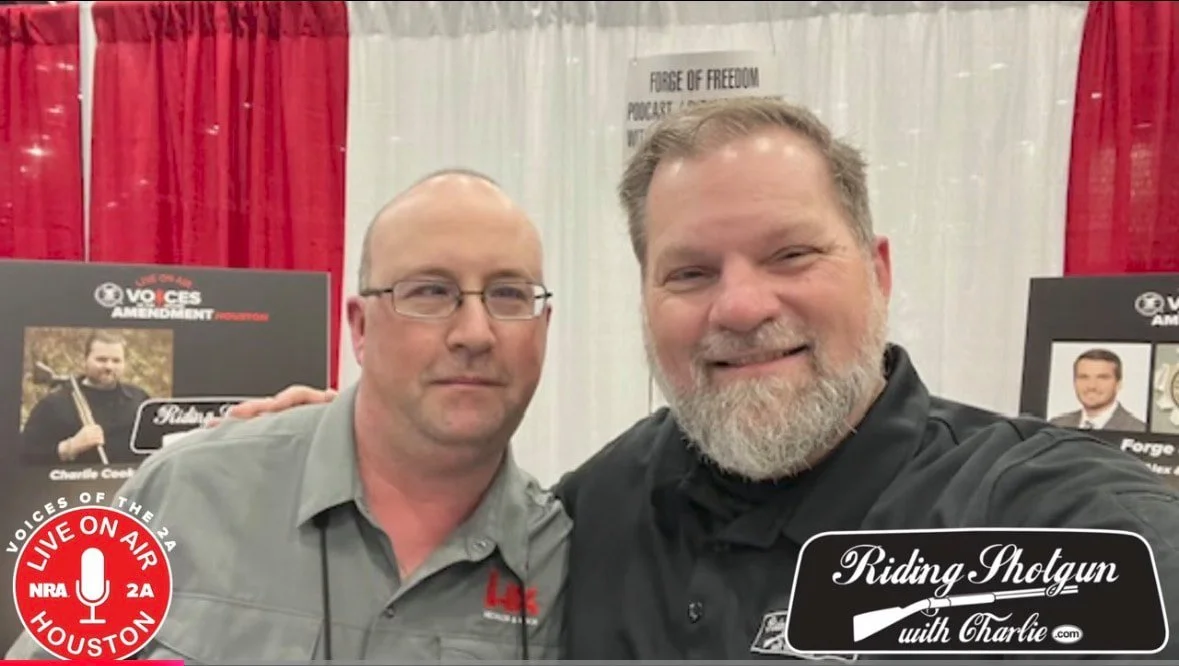 Two men taking a selfie at an event with posters in the background. One man has a beard and the other wears glasses. There are signs that include the phrases 'Riding Shotgun with Charlie' and 'Voices of the 2A'.