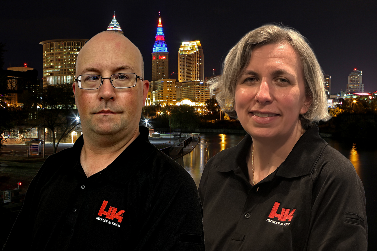 A man and a woman wearing black shirts with a red and white logo, standing outdoors at night with a city skyline and illuminated buildings in the background.