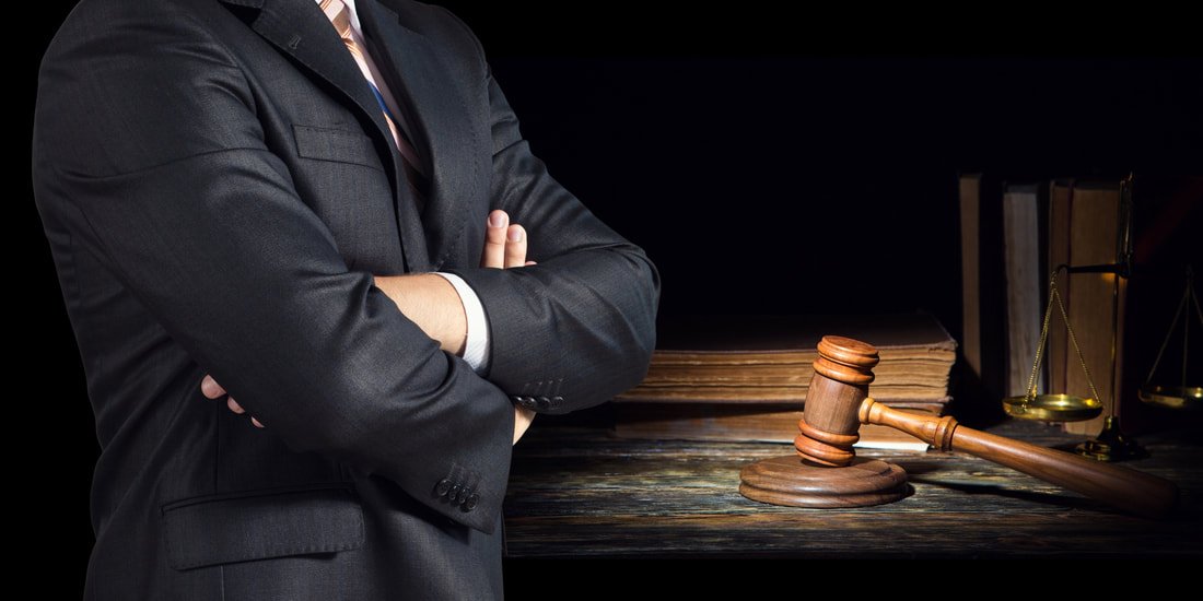 A person in a suit standing with arms crossed, on a wooden desk with a judge's gavel, legal books, and scales of justice.