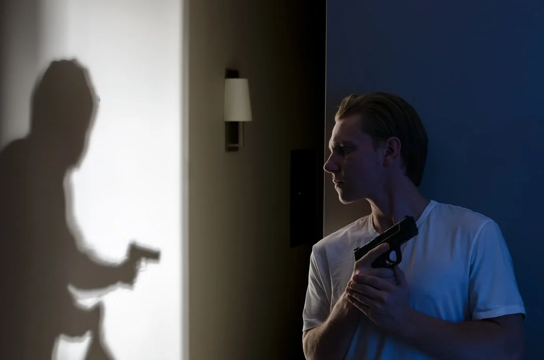 A young man with a gun in his hand, standing in a dimly lit room, facing a wall where his shadow is cast. The shadow on the wall depicts a man holding a gun and reading a paper or a card.