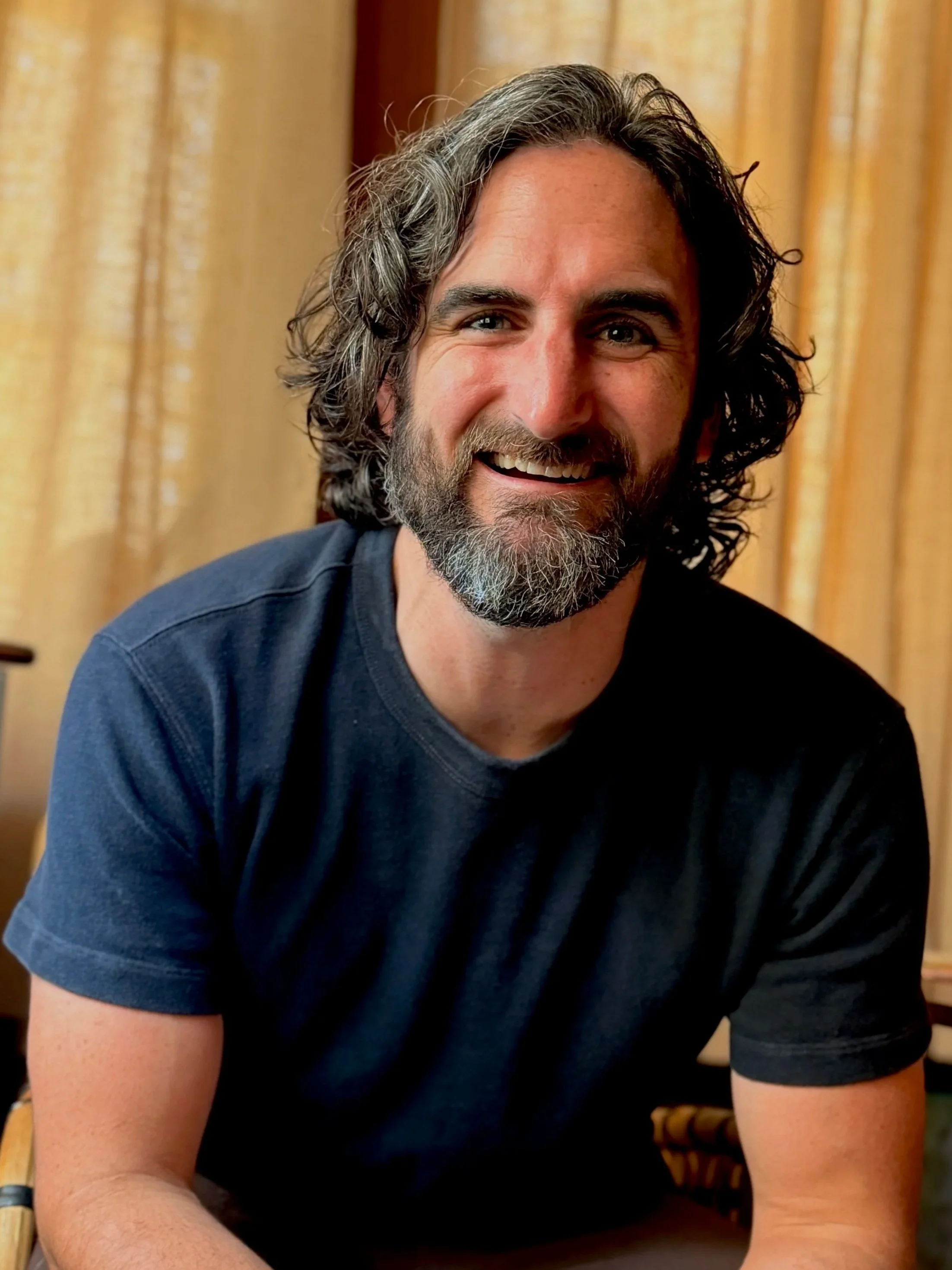 A man with long, wavy gray hair and a beard, smiling, wearing a dark shirt, sitting in front of beige curtains.