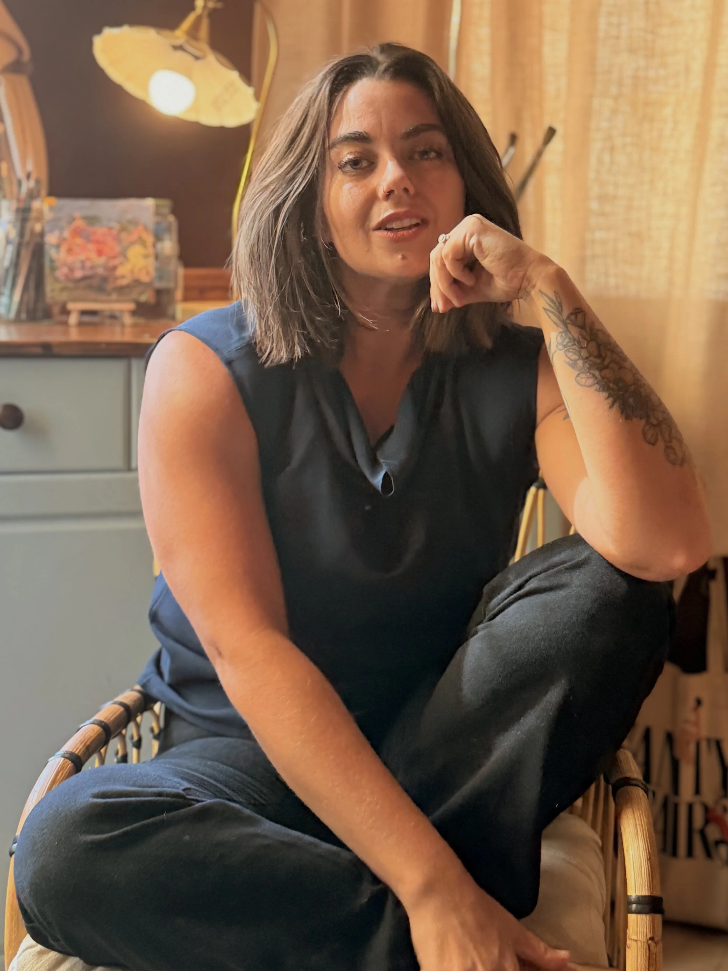 Woman with shoulder-length brown hair, wearing a black top and black pants, sitting in a wicker chair, with tattoo on her left forearm, in a room with beige curtains and a lamp in the background.