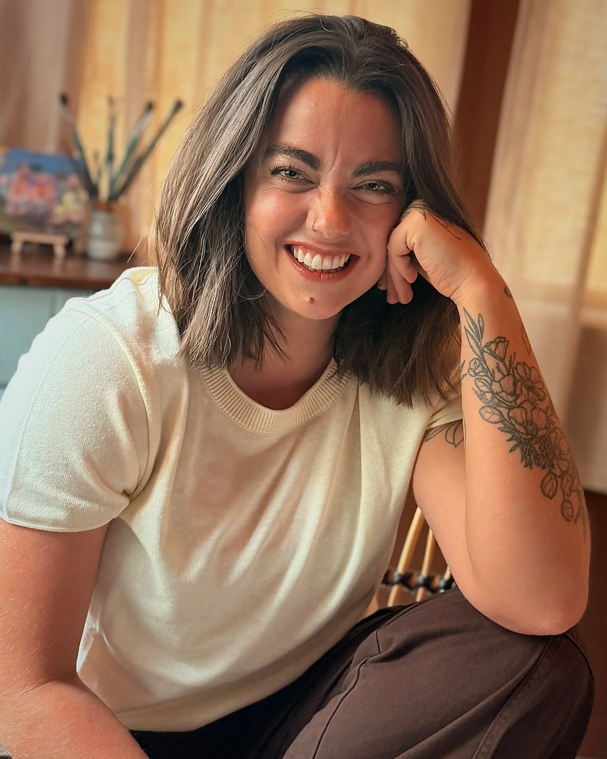 A smiling woman with long dark hair, wearing a nose ring and visible tattoos on her right arm, sitting with her hand resting on her cheek in a cozy indoor setting with curtains in the background.