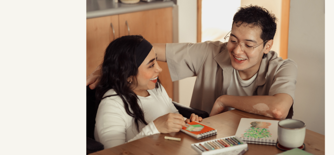 A young woman with dark hair, wearing a white shirt and a black headband, sitting at a table with a man with glasses, short dark hair, and an arm with a skin condition. They are smiling at each other. On the table are art supplies, a drawing, and a coffee mug.