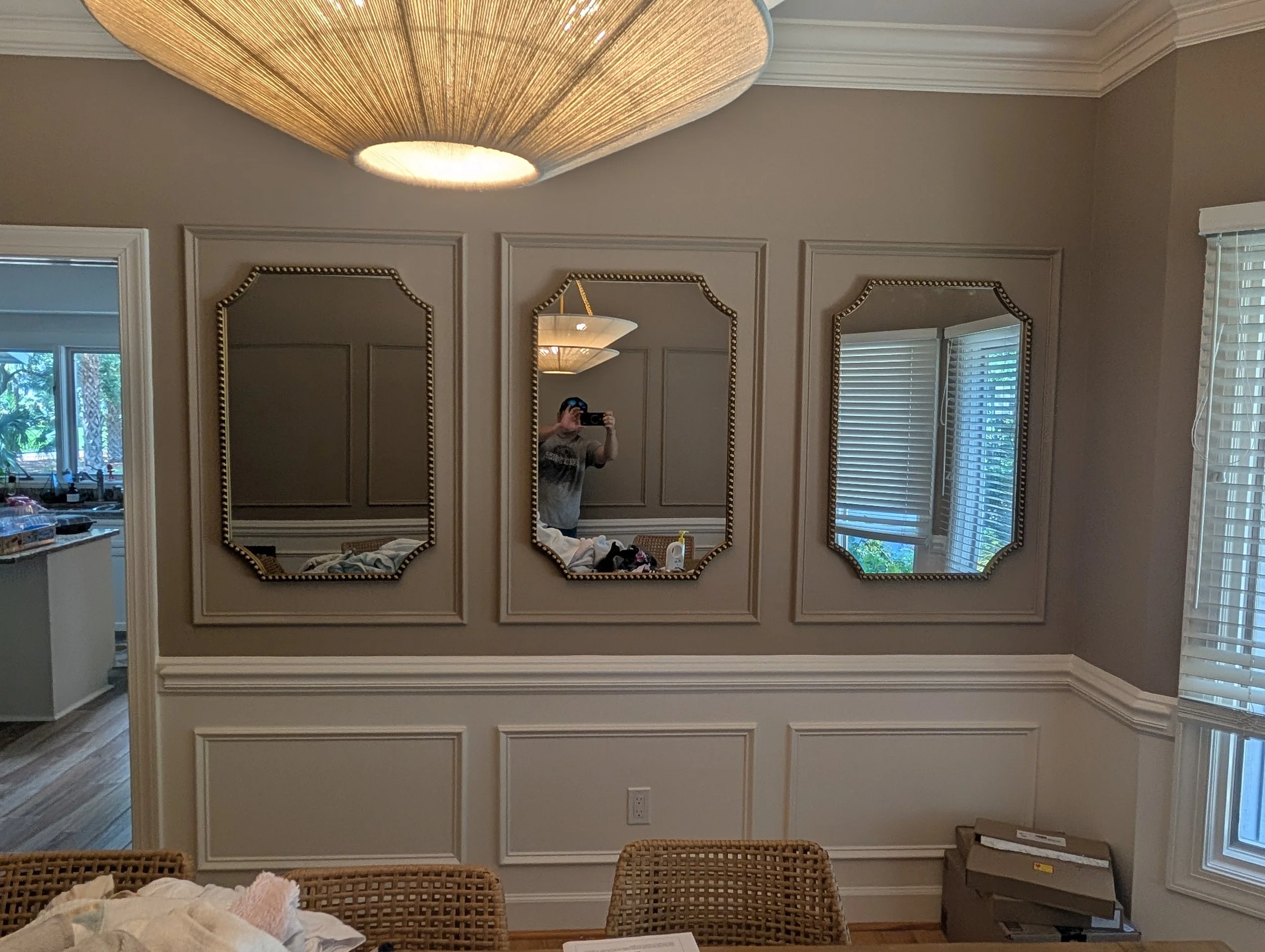 A wall with three framed mirrors reflecting a person taking the photo, near a window with blinds, and a ceiling light fixture.