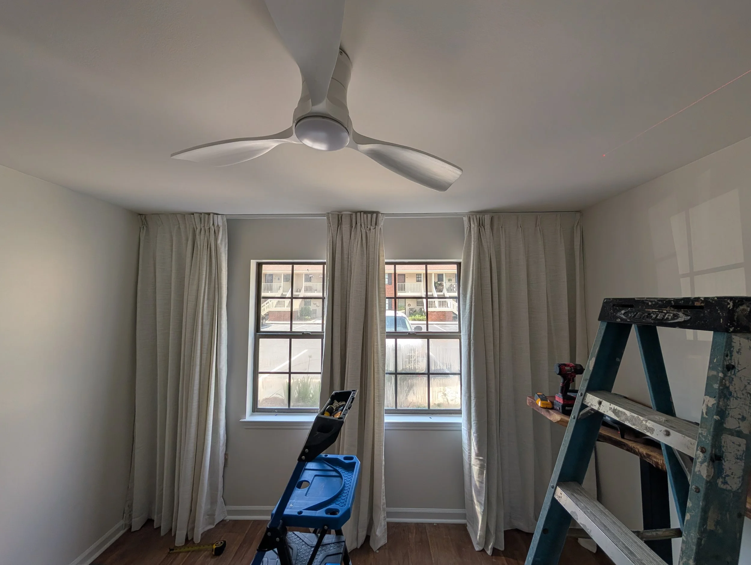 A room with two large windows with beige curtains, a white ceiling fan, a ladder, and a small blue step stool with tools on it, indicating renovation work.