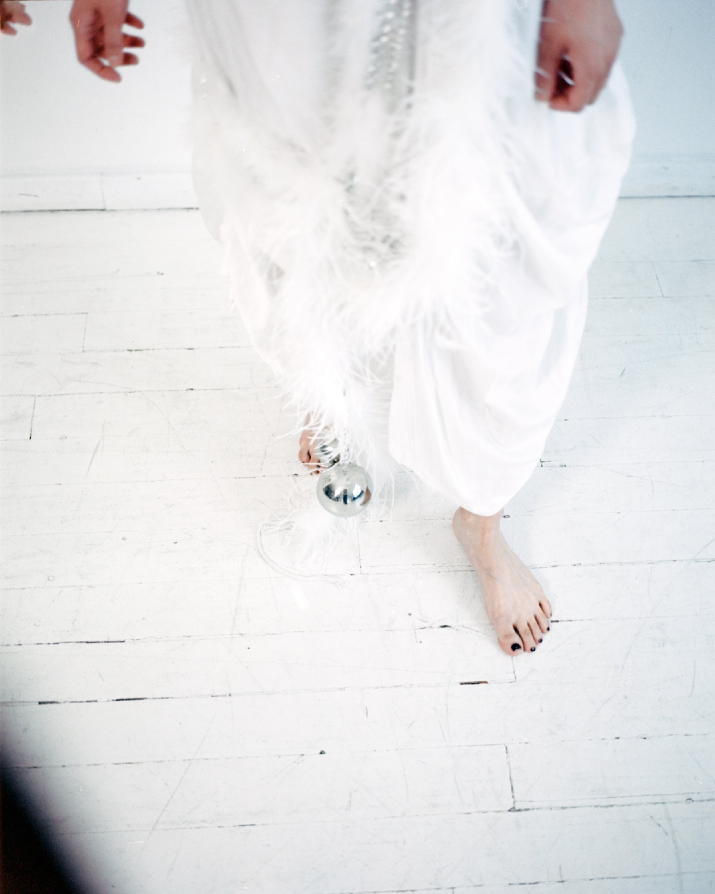 A person wearing a white dress and black nail polish is taking a mirror selfie. The person is standing on a white wooden floor, holding a shiny silver ball in their right hand, with their face not visible. Their toes are painted black.