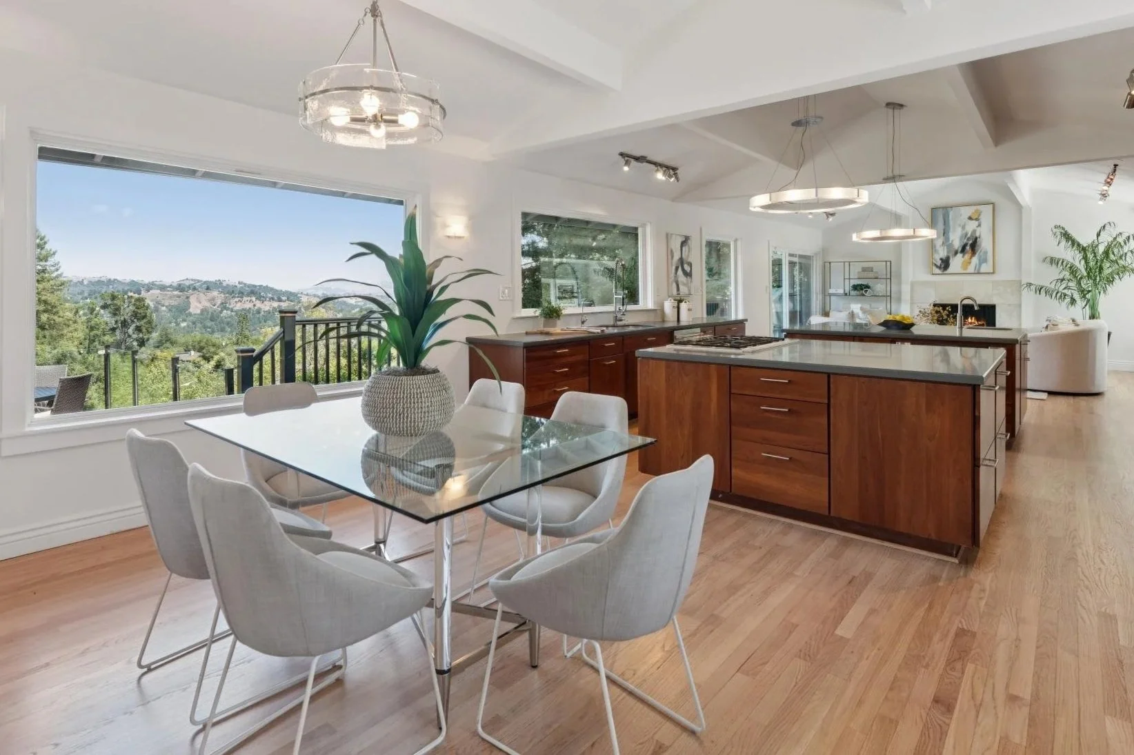 Interior view of a modern, open-concept kitchen and living room with large window showing scenic outdoor landscape. The kitchen features wooden cabinets, an island with a white countertop, and modern lighting fixtures. A dining table with seven white upholstered chairs and a decorative plant in a gray pot are in the foreground.
