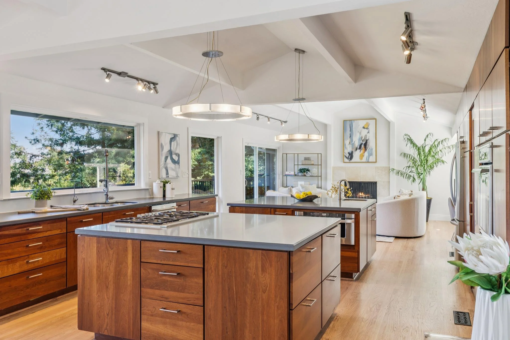 A modern kitchen with white walls and ceiling, wood cabinets, a kitchen island with a white countertop, large windows showing trees outside, and contemporary lighting fixtures.
