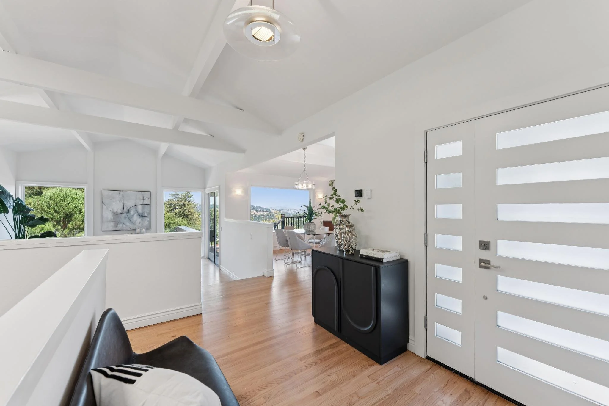 Modern white living room with large windows, hardwood floors, black armchair with a pillow, and decorative vases on a black cabinet near a white door with rectangular windows.