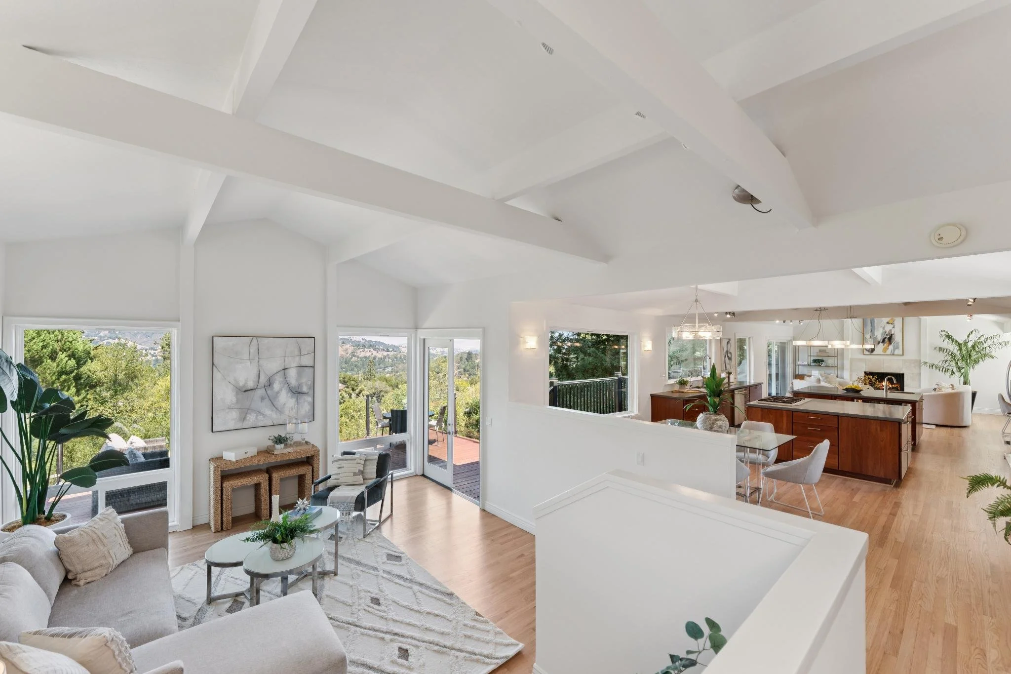 Open-concept living room and kitchen with white walls, large windows, and modern decor, overlooking a green outdoor landscape.