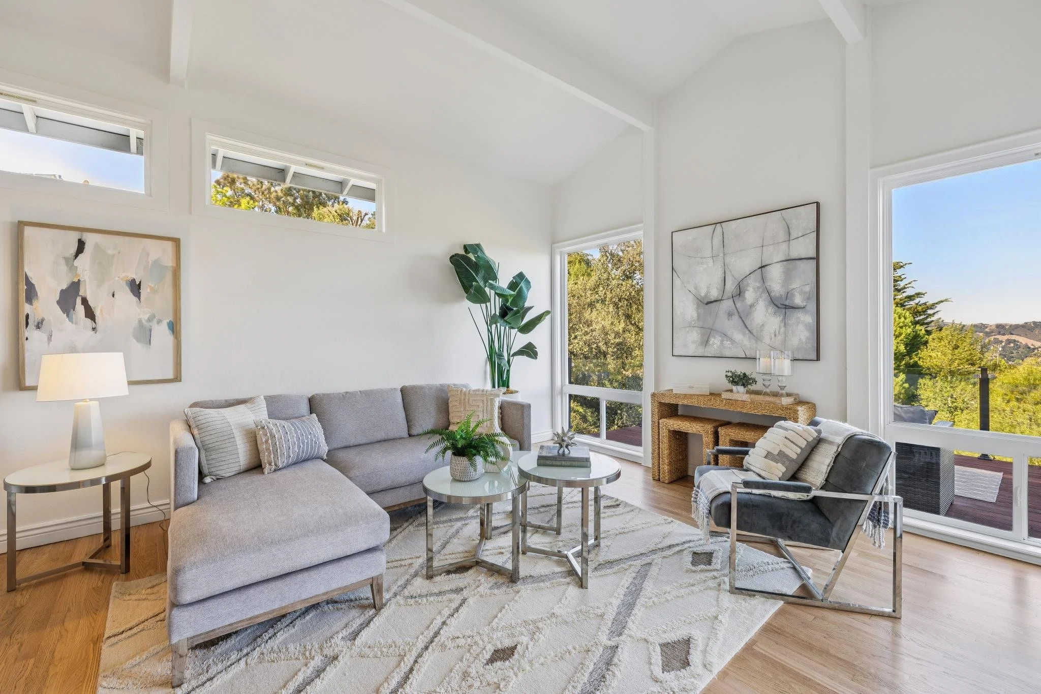 Bright living room with large windows, gray sofa, patterned pillows, white side table with lamp, abstract artwork, green plants, and a fireplace, with a view of trees outside.