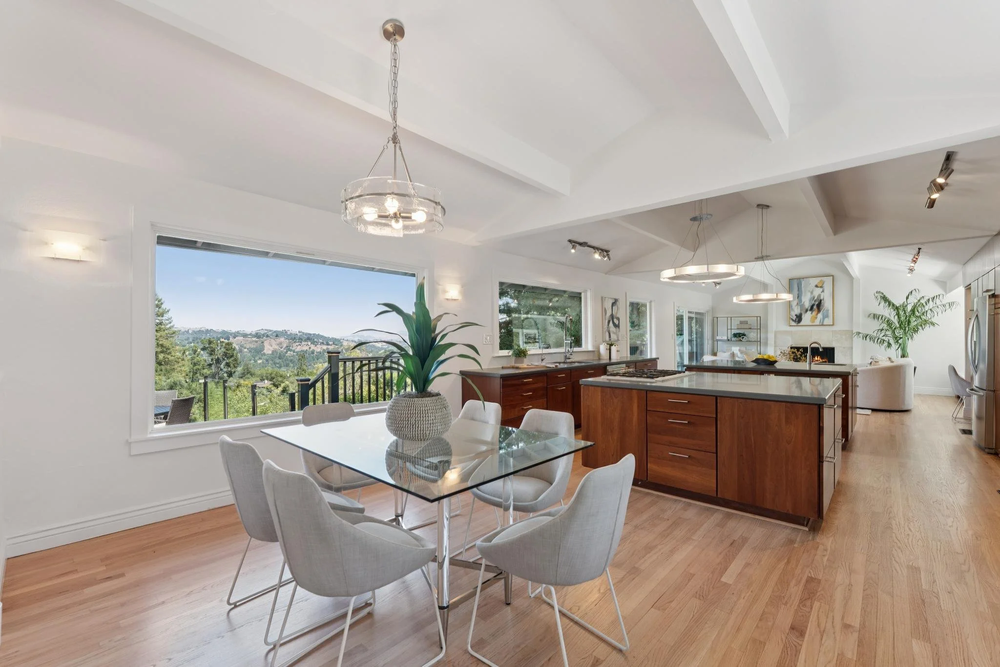 Modern living space with large window, white walls, wooden floor, glass dining table with white chairs, kitchen island, abstract wall art, plants, and various lighting fixtures.