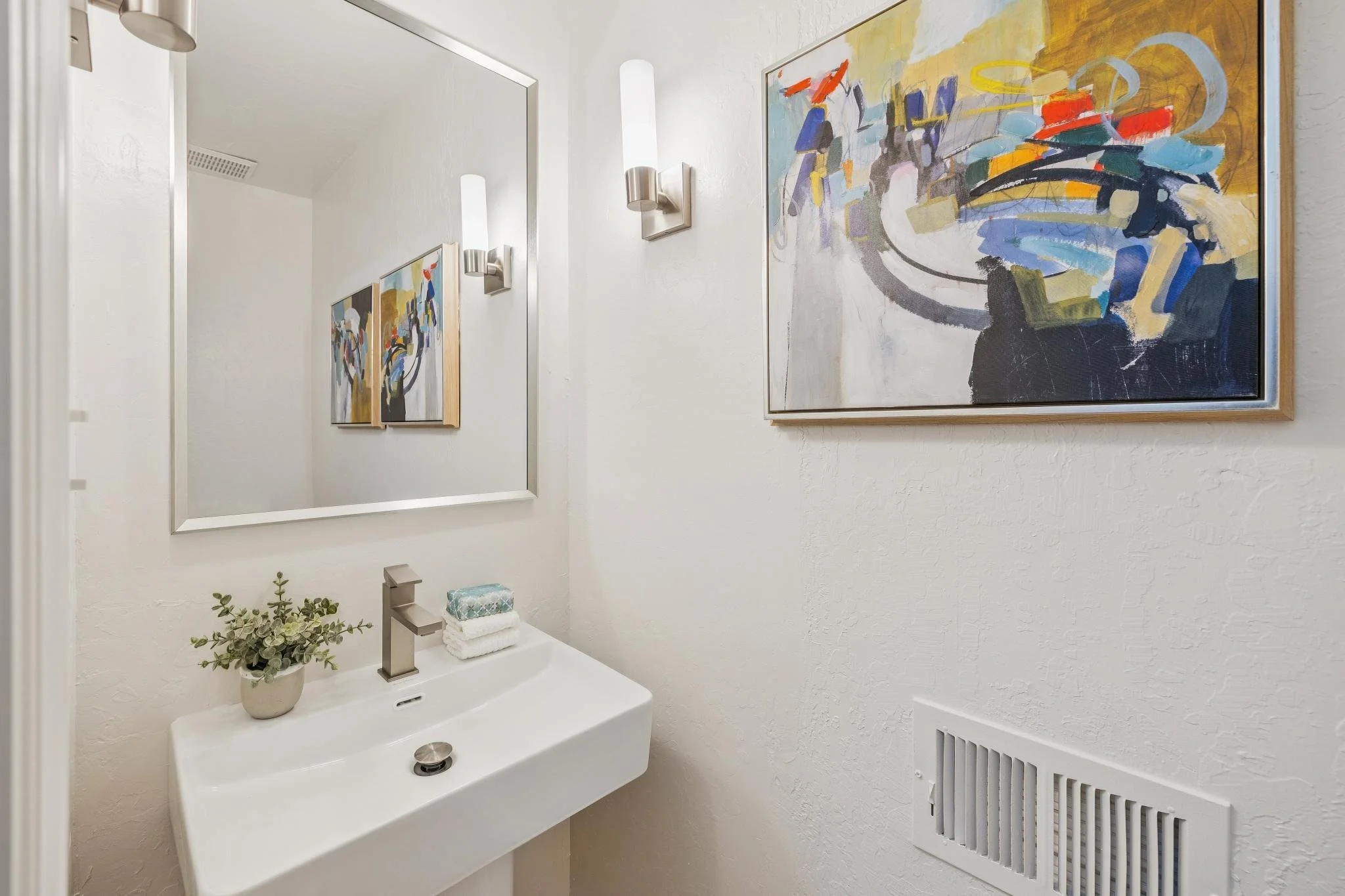 A bathroom with a white sink, a large mirror, a wall-mounted light fixture, abstract artwork on the wall, a small plant, and a stack of folded towels.