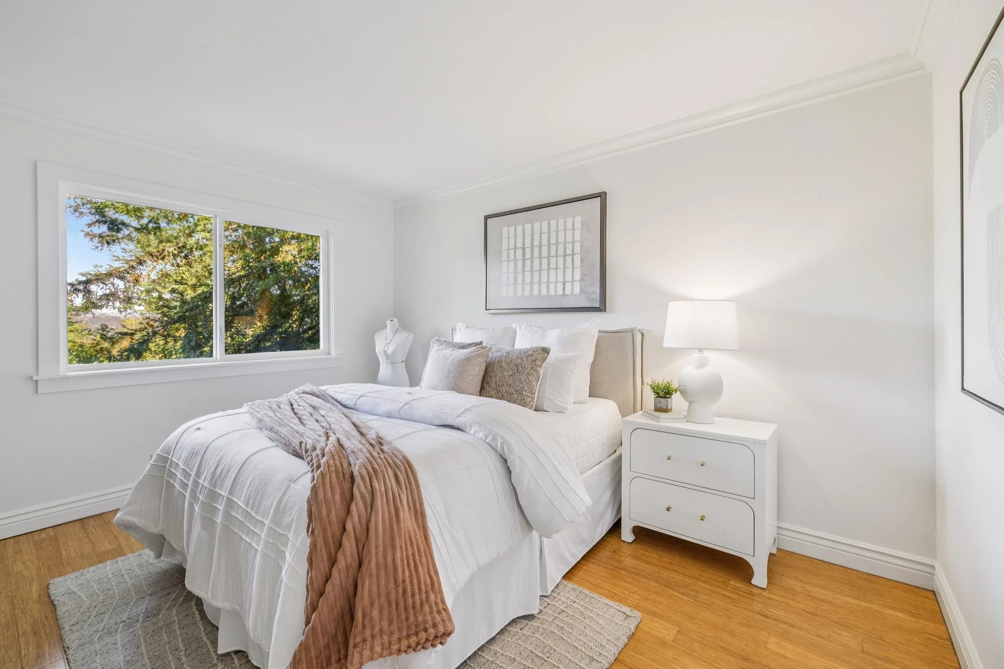 A bedroom with a large window showing green trees outside. The bed has white bedding, pillows, and a rust-colored throw blanket. There's a white bedside table with a white lamp and small plant. Artwork hangs on the wall above the bed.