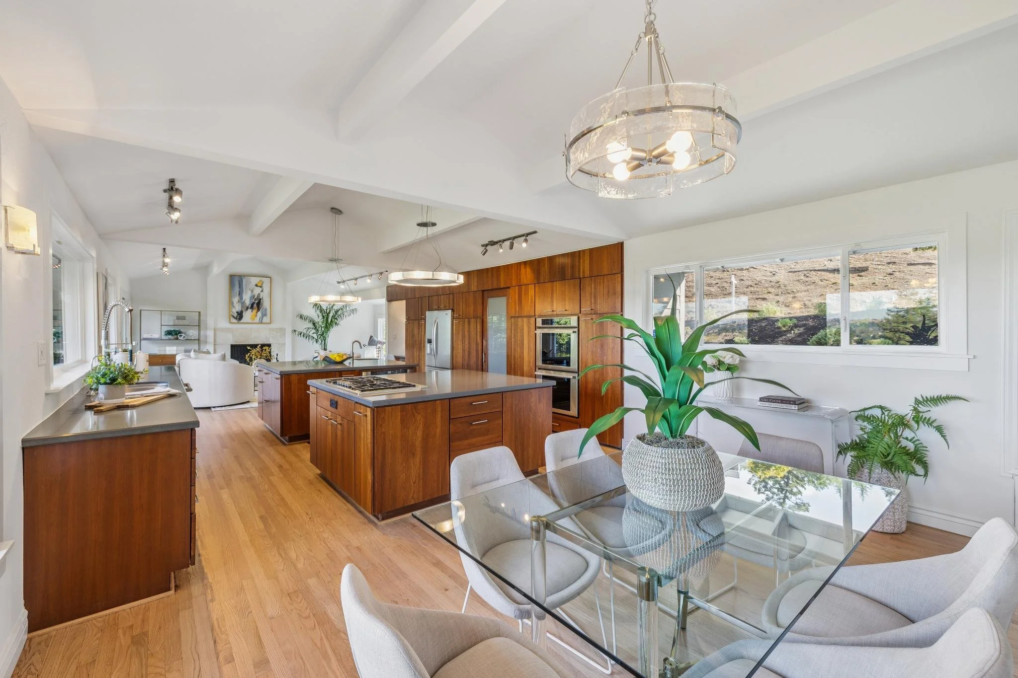 Open-concept kitchen and dining area with wooden cabinets, large windows, and modern lighting fixtures. There are plants on the table and near the windows, with a view of the outdoors through the windows.