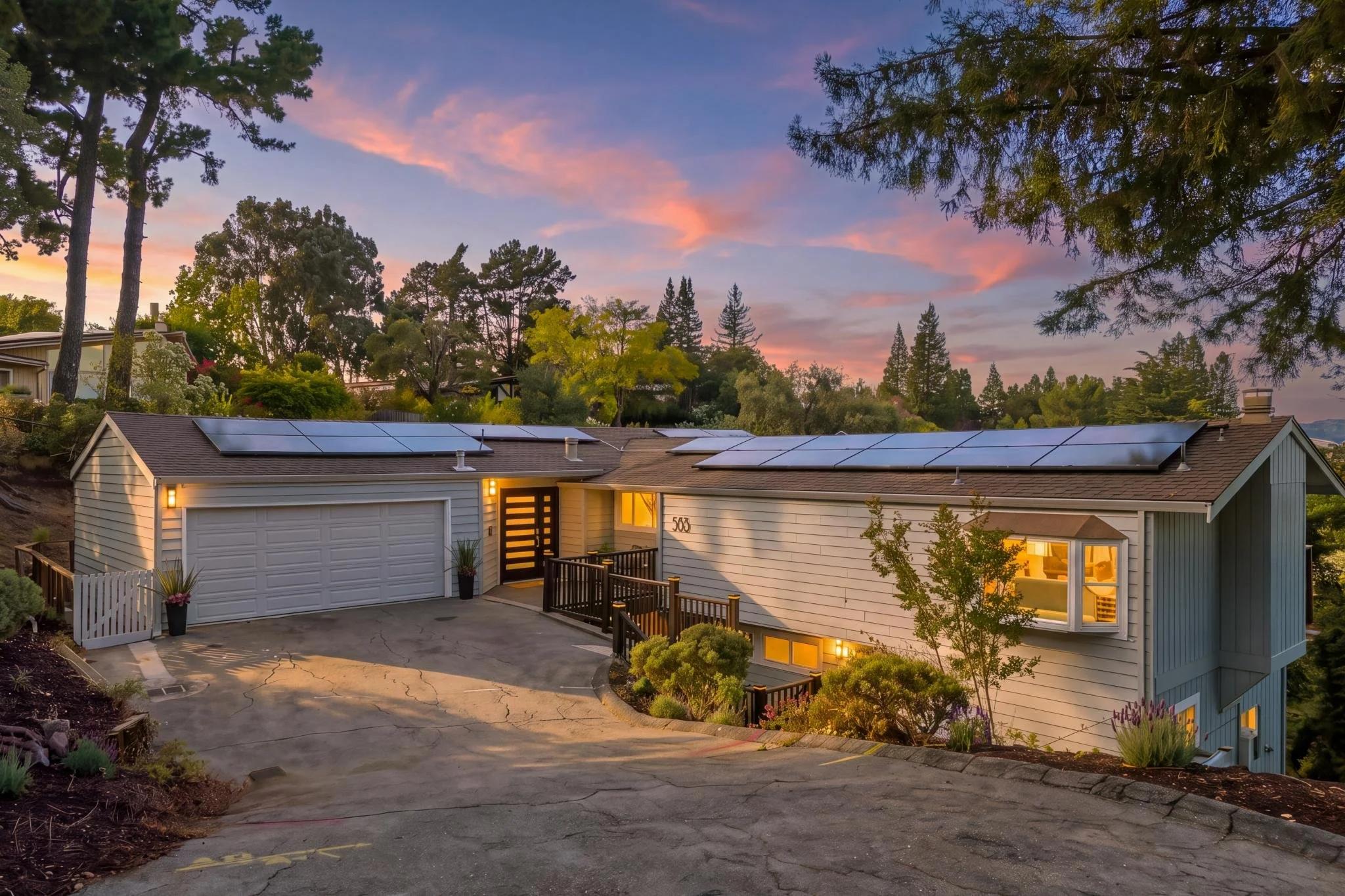 A house with solar panels on the roof, situated in a lush green neighborhood at sunset. The sky has pink and purple clouds, and the house has warm lighting inside. There is a driveway with some plants and flowers near the entrance.