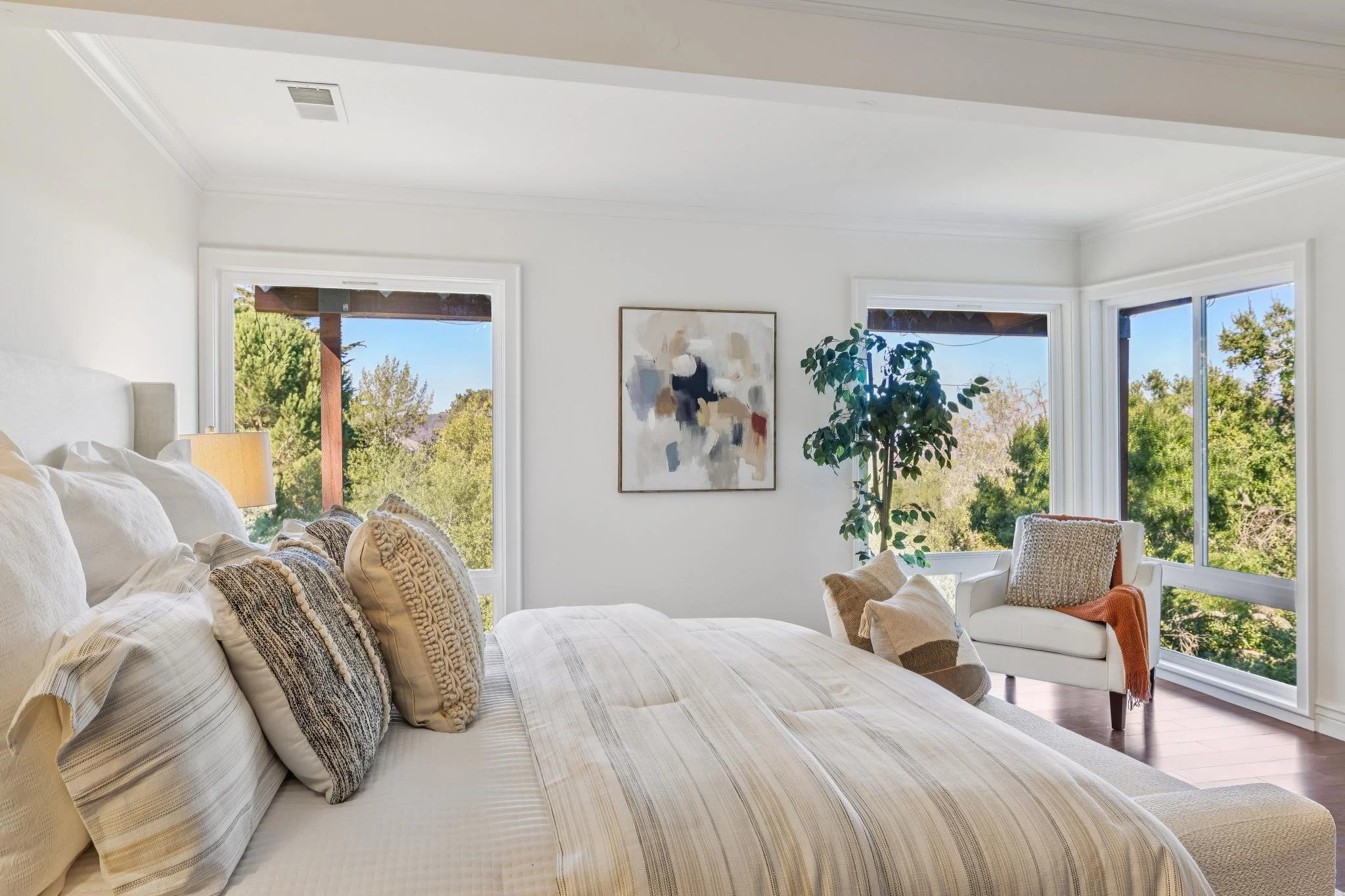 A bright, airy living room with white walls and large windows showcasing trees outside. The room features a white sofa with various textured throw pillows, a white armchair with an orange throw blanket, a tall green houseplant, and a modern abstract 