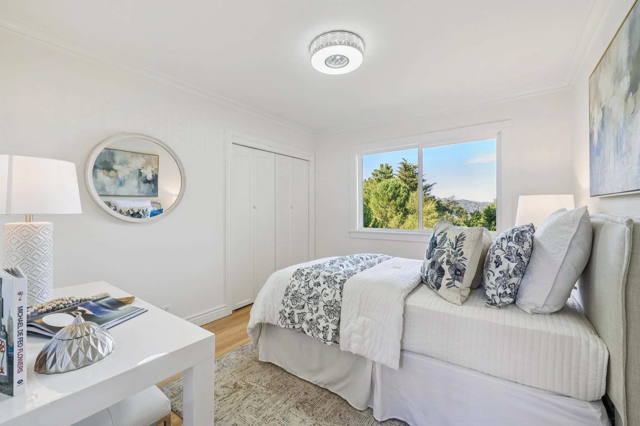 Bright bedroom with white walls, large window showing trees and sky, white bed with floral patterned pillows and cover, round mirror on the wall, white lamps on bedside tables, abstract artwork, and a closet with sliding doors.