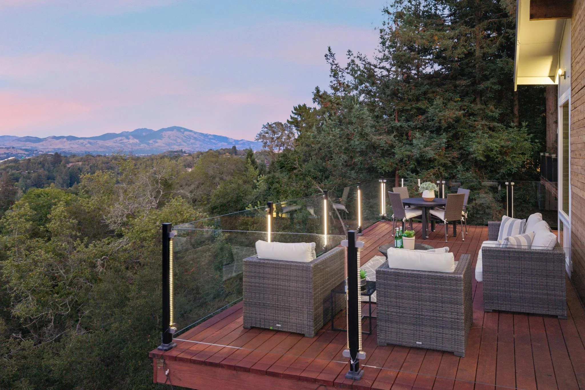 A backyard deck with outdoor furniture, overlooking a tree-filled landscape with mountains in the distance during dusk.