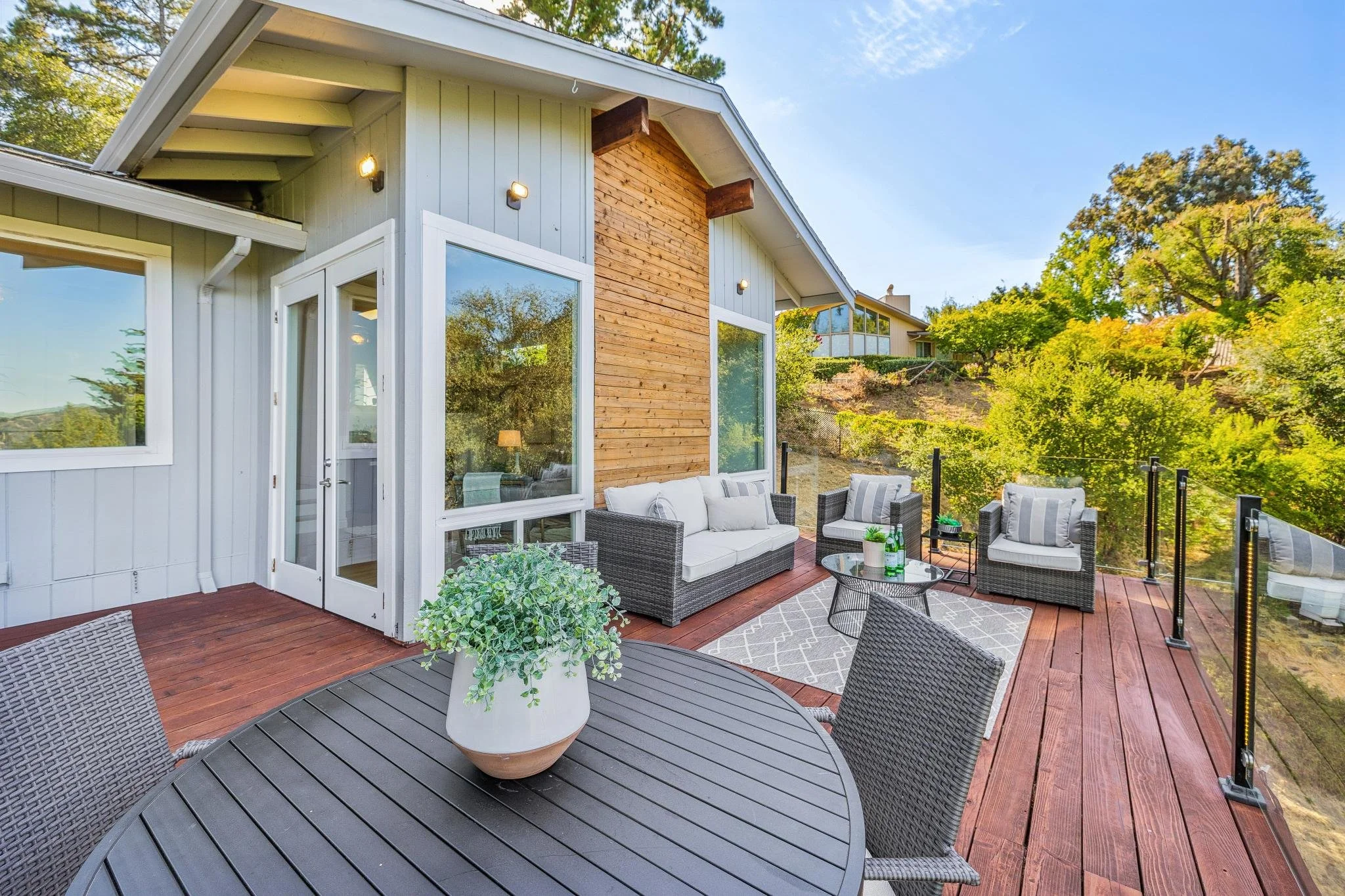 Outdoor patio area with seating, including a gray wicker sofa, chairs, a round table, and green potted plants, overlooking trees and neighboring houses.