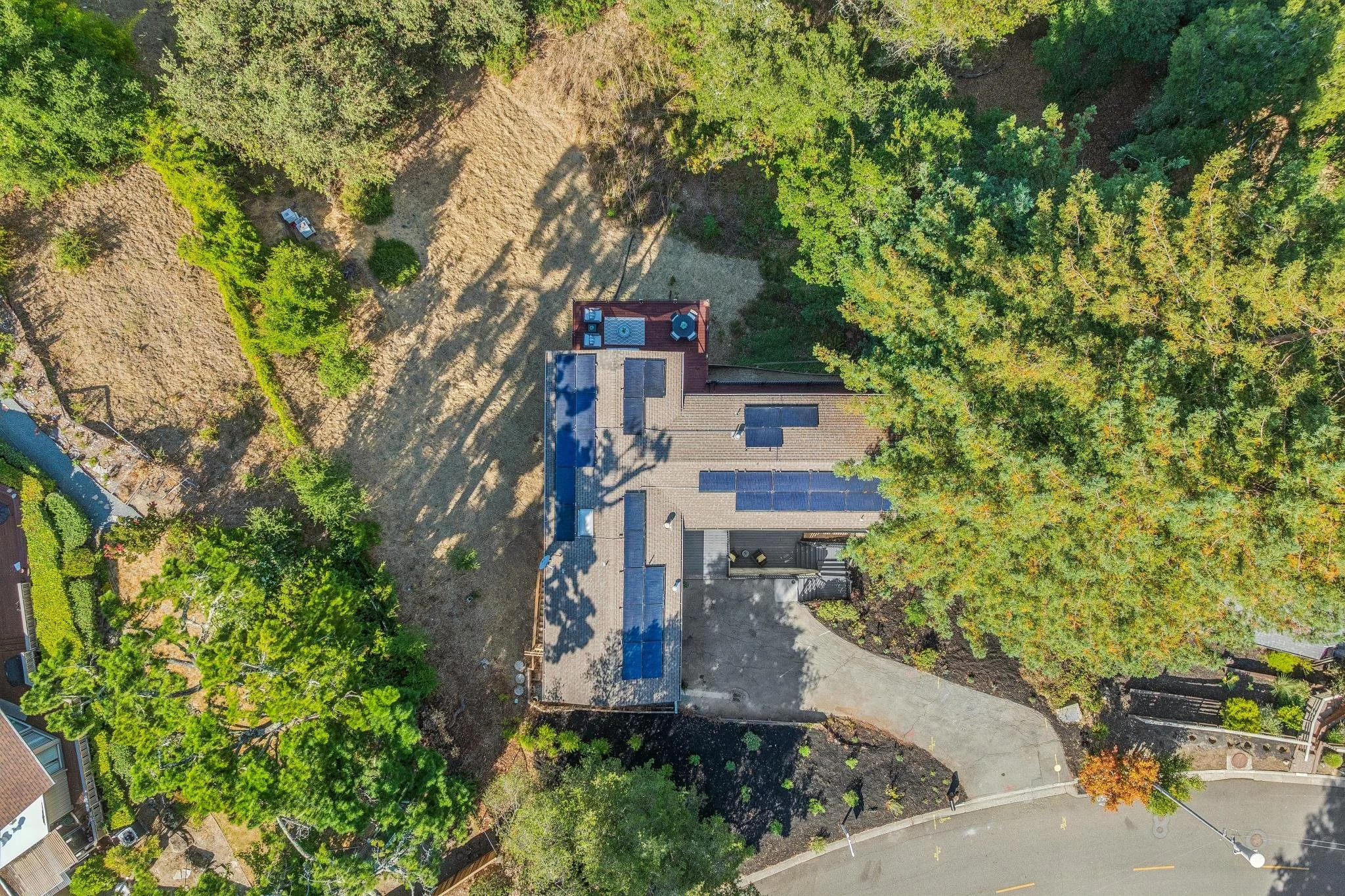 An aerial view of a residential backyard with trees, solar panels on the roof, and outdoor furniture on a deck.