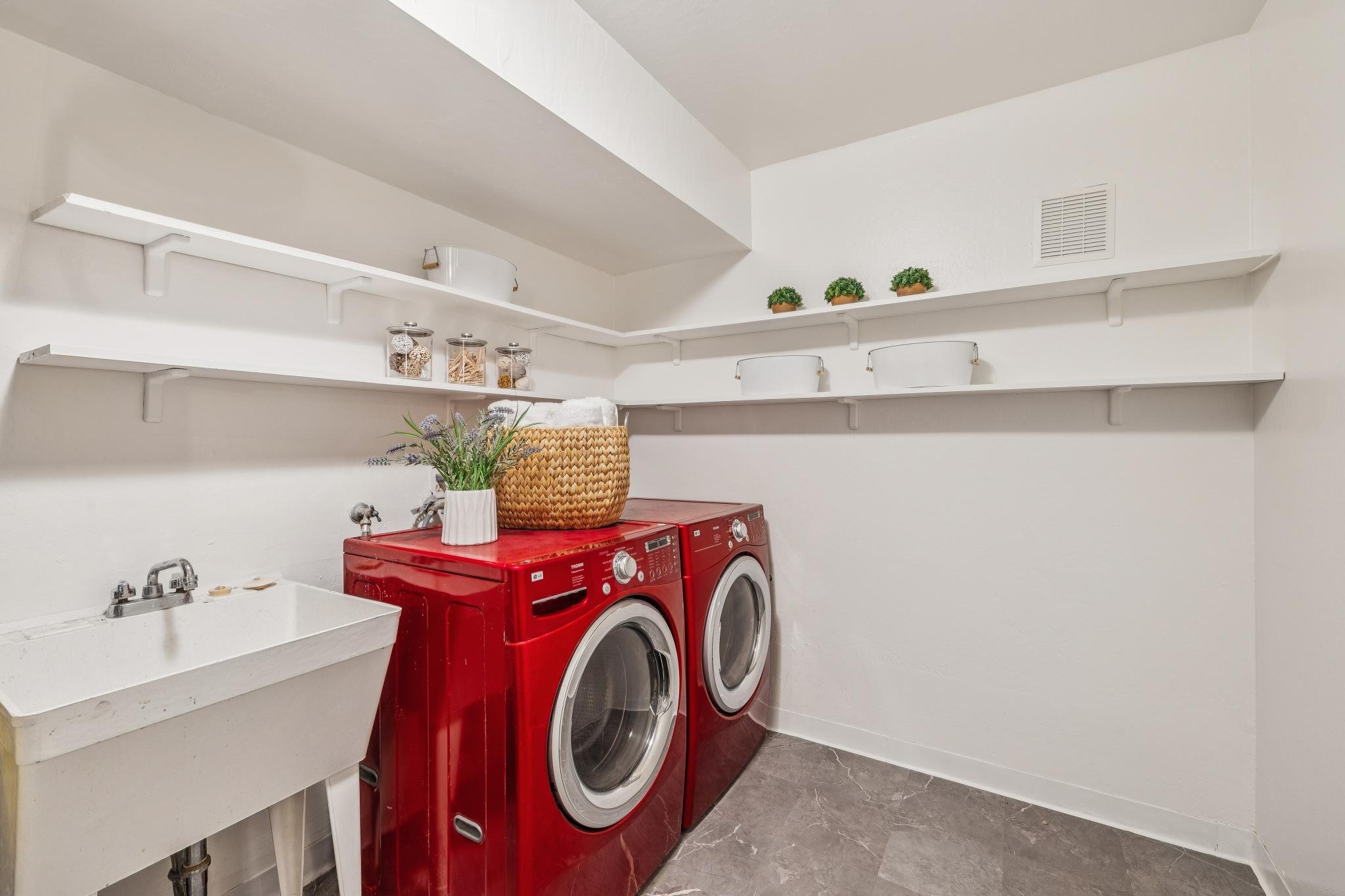 A laundry room with two red front-loading washing machines side by side. Above them are two white shelves with small potted plants and jars of laundry essentials. To the left, there is a utility sink, and on the countertop, there is a white vase with