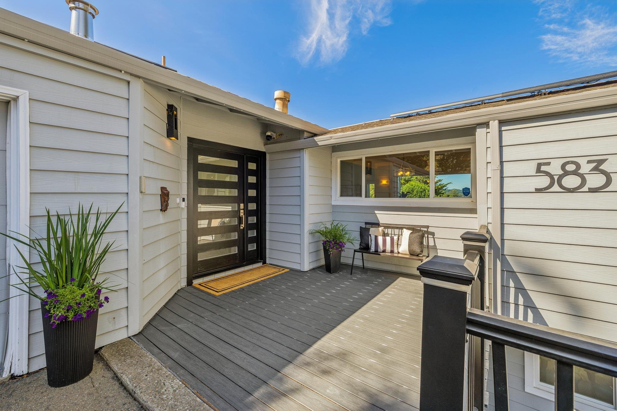 Front porch of a house with a gray wooden floor, black front door, a white wall with the house number 583, a window, potted plants, and outdoor furniture under a blue sky.