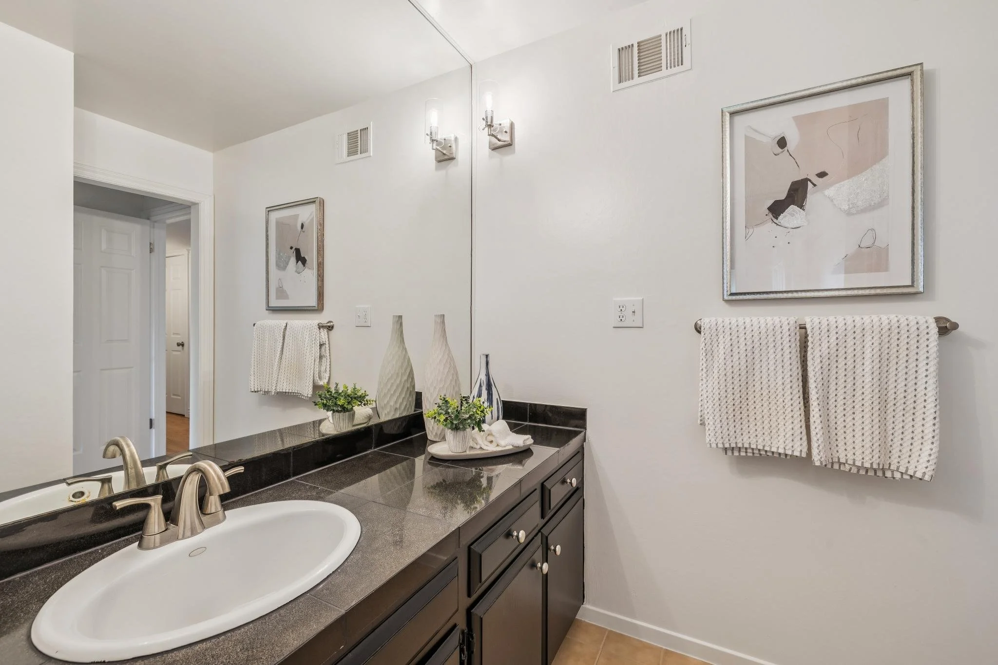 Bathroom with white walls, two framed abstract artworks, two wall-mounted light fixtures, a large mirror, dark vanity with multiple drawers, a white oval sink, silver faucets, decorative vases, a potted plant, and towel racks with towels. Ventilation