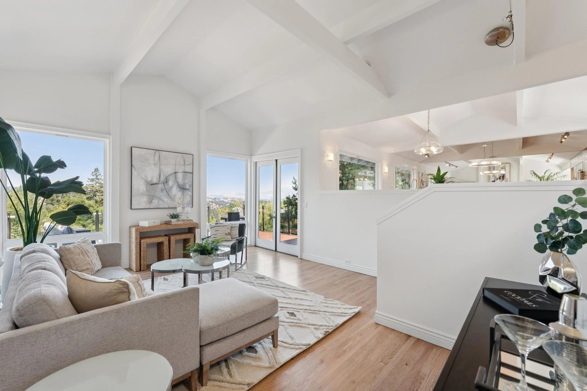 Bright living room with white walls, large windows, a beige sofa, glass coffee tables, indoor plants, and a view of trees outside.