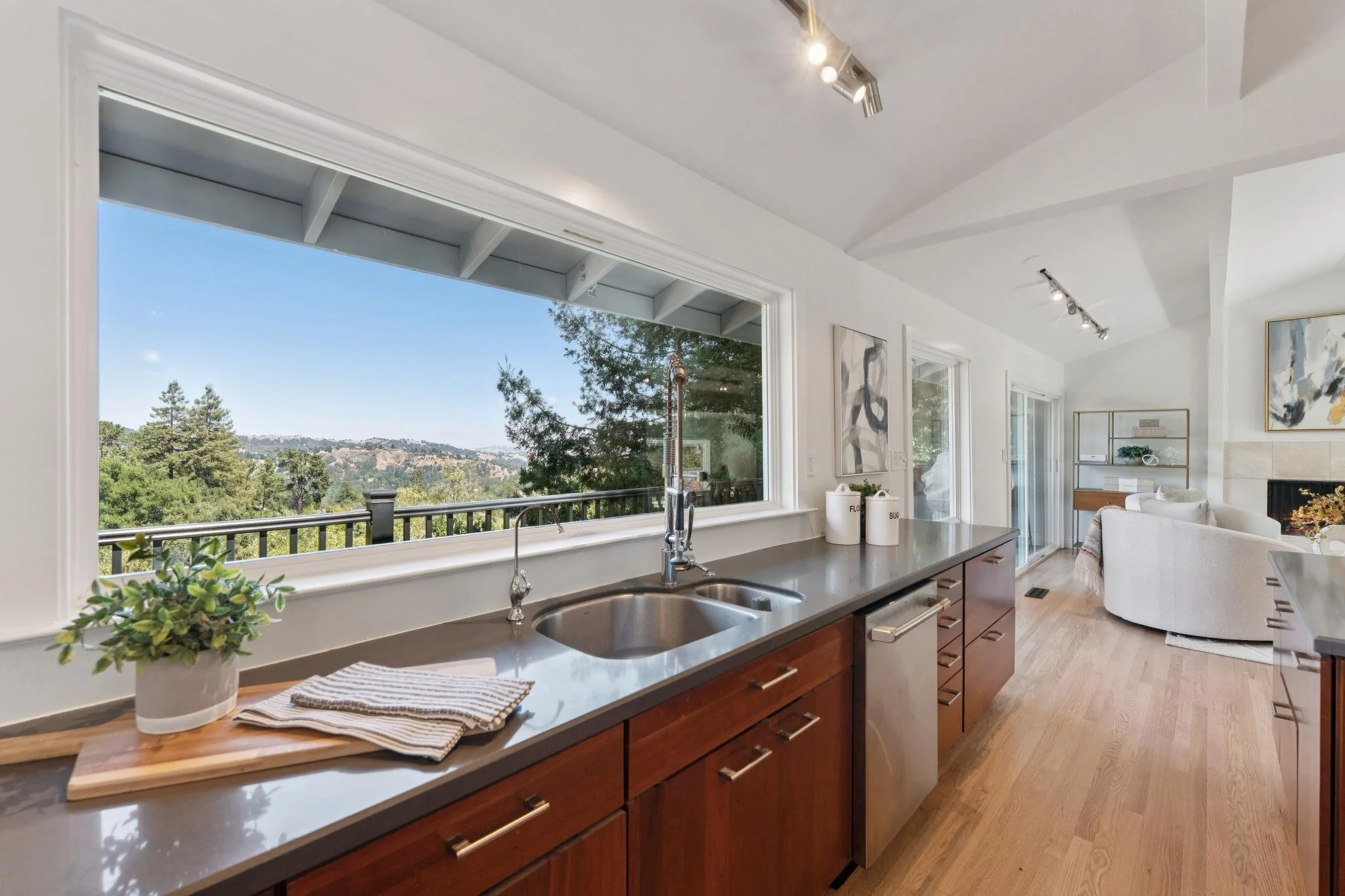 View of a modern kitchen with a large window overlooking green trees and hills, a double sink, and wooden cabinets, with a living room area visible in the background.