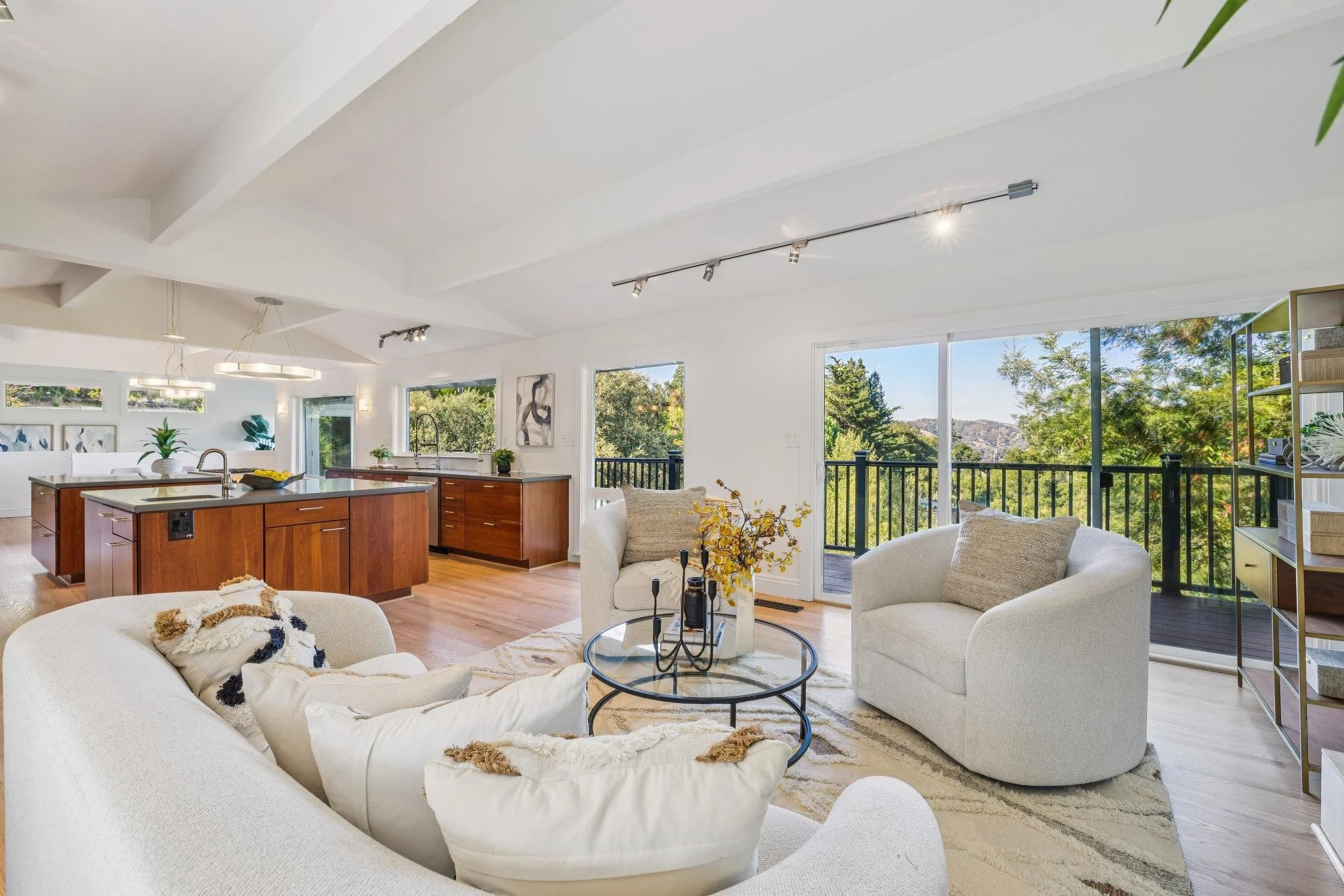 Bright, modern living room with white walls, large windows showing trees outside, a white sofa, chairs, a round glass coffee table, and a kitchen area with wooden cabinets.