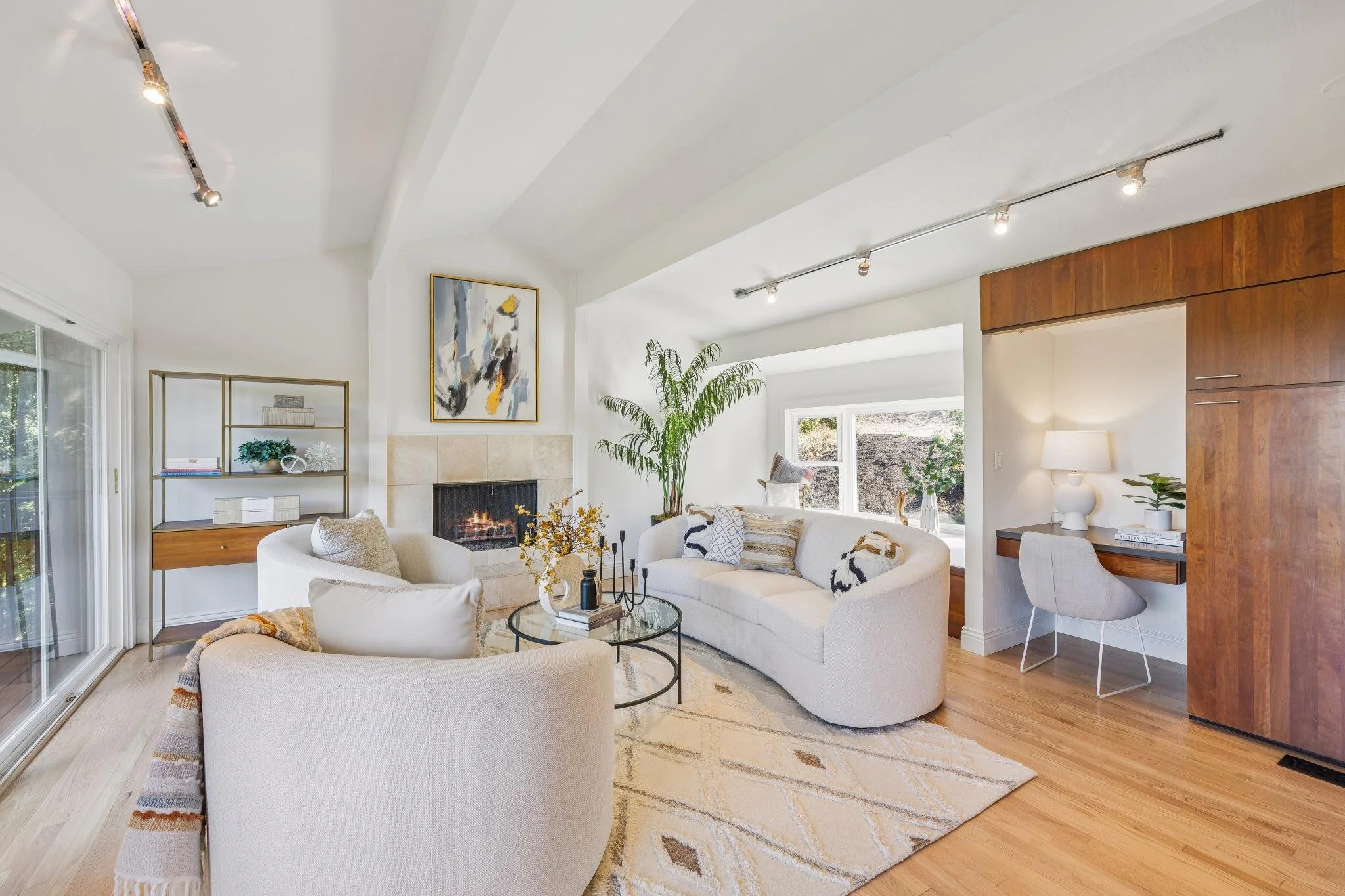 Living room with white curved sofa, fireplace, large window, houseplant, abstract art on wall, and wooden accents.