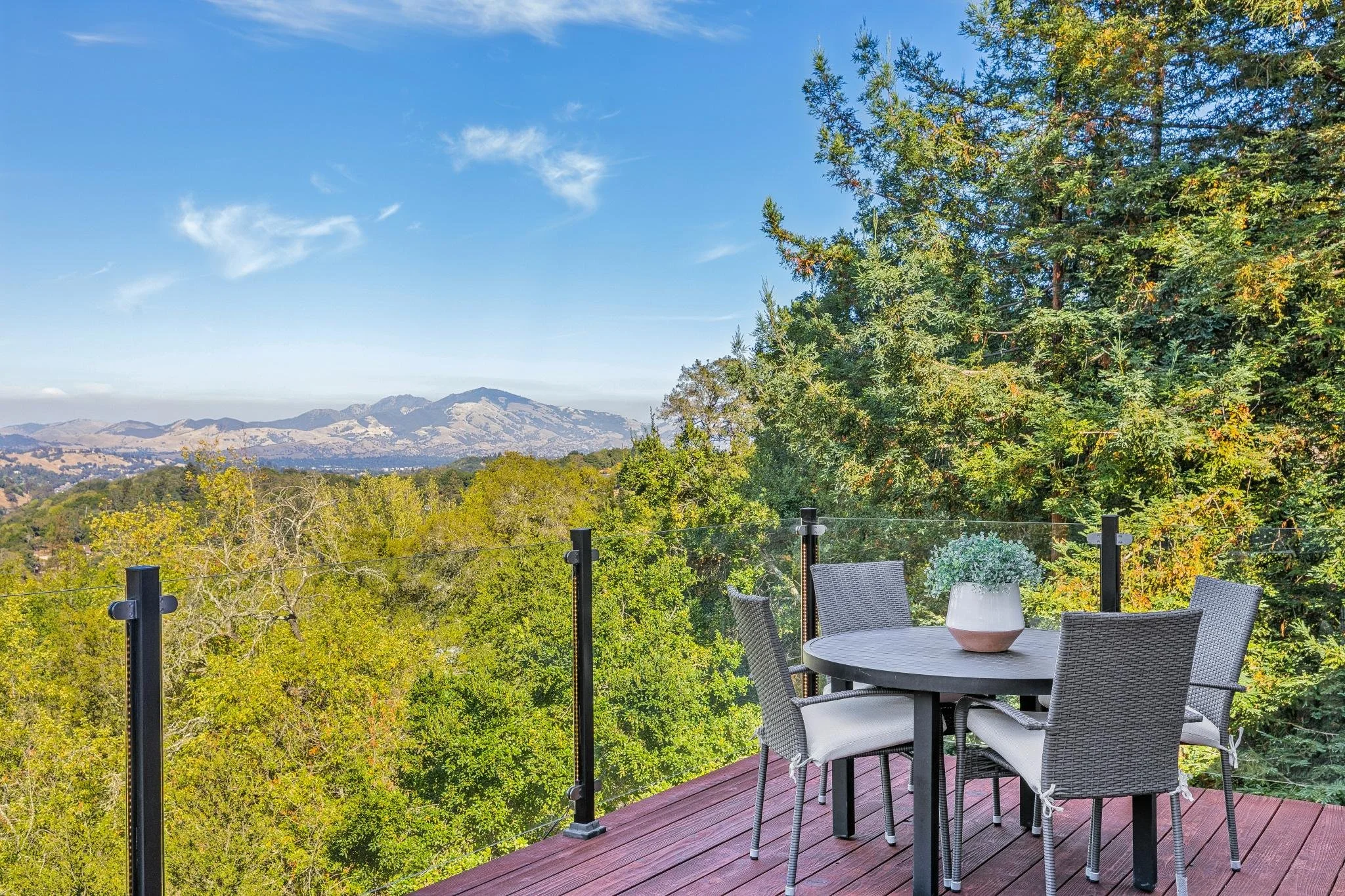 A patio with a round table and four chairs, a potted plant on the table, and a view of trees and mountains in the distance under a partly cloudy sky.