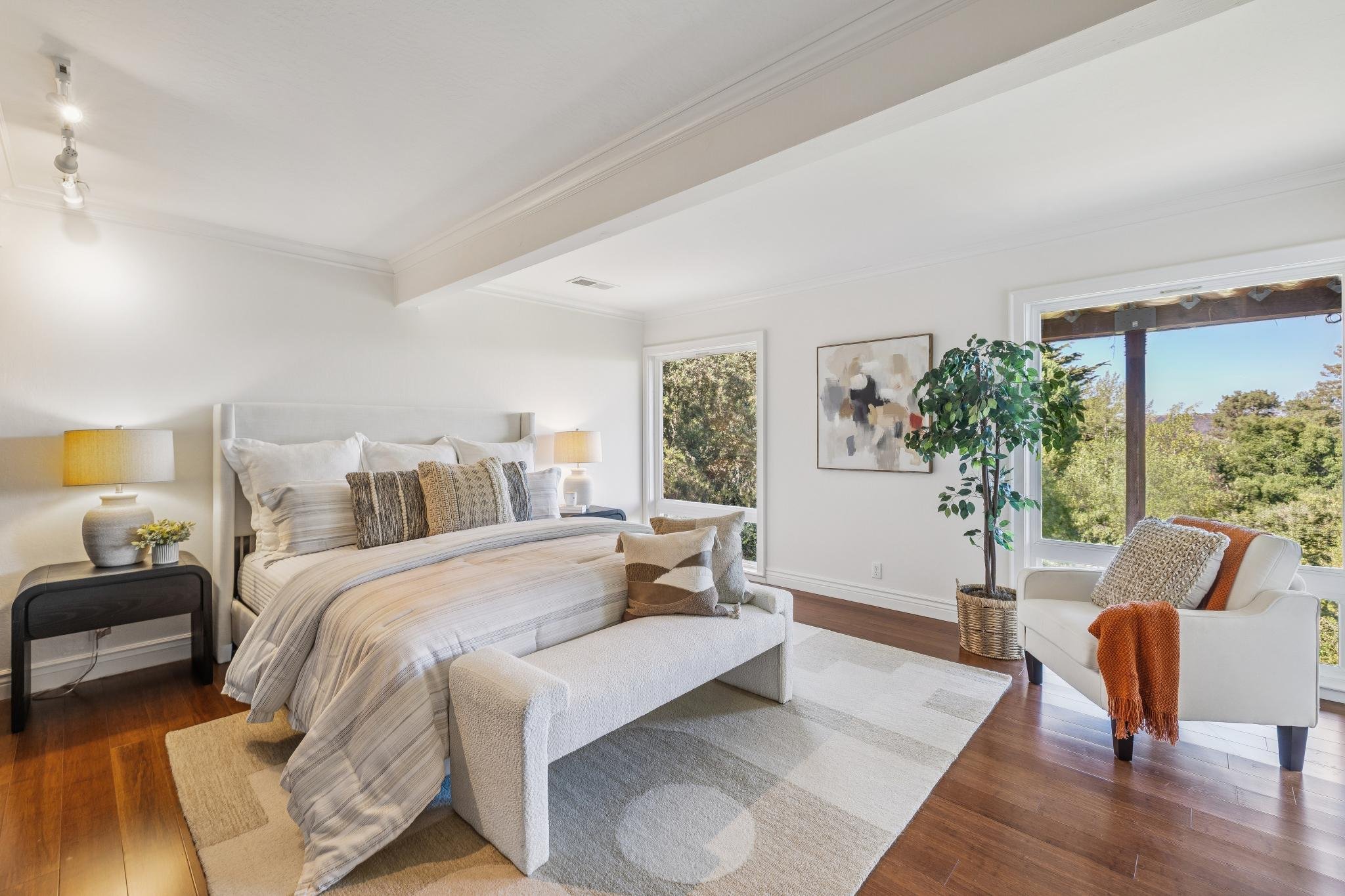 Bright bedroom with white walls, large windows showing trees outside, a bed with striped bedding, matching pillows, and white upholstered headboard. A black nightstand with a yellow lamp, a small plant, and decorative object. Artwork on the wall, a white armchair with an orange throw, a potted plant, and wooden flooring with a beige rug.